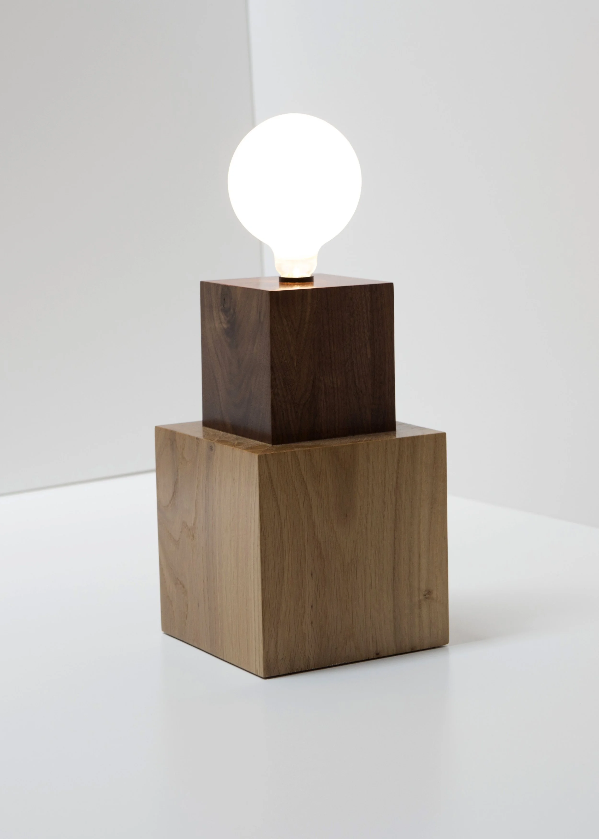 A modern table lamp with a white spherical bulb on a dark wooden block, placed on a light wooden pedestal against a plain light-colored background.