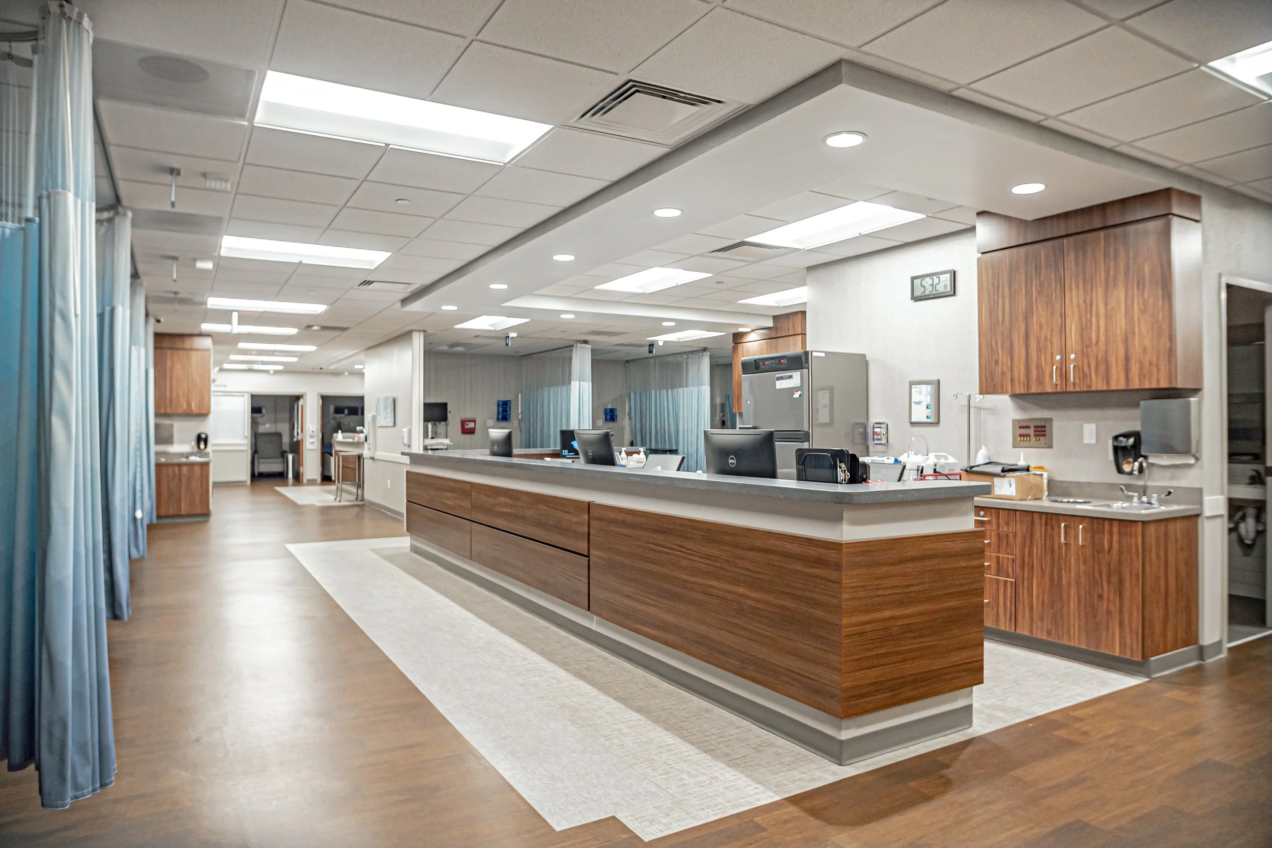 Hospital reception area with a curved wooden counter, computers, medical equipment, and curtains separating patient spaces.