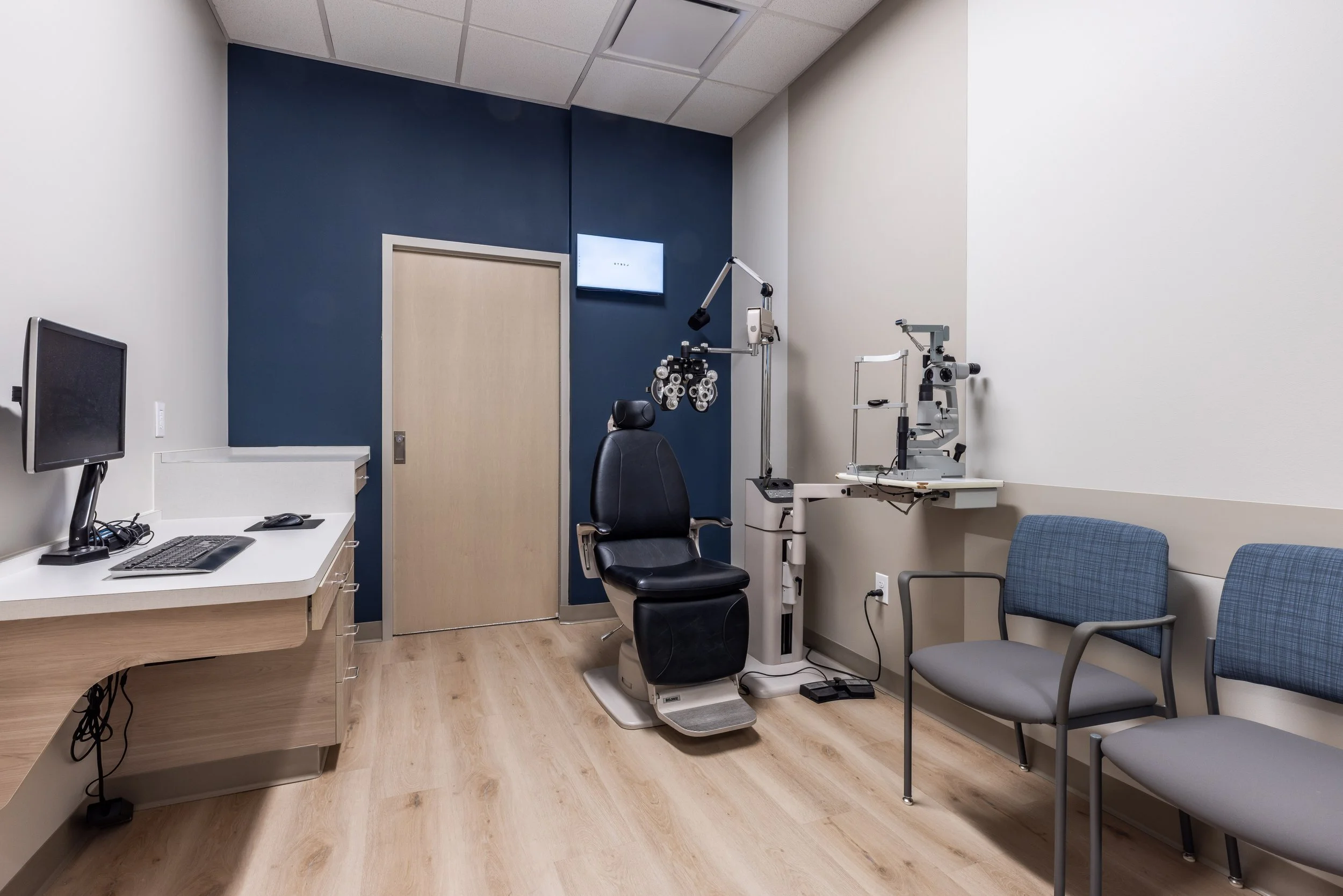 Medical exam room with an eye examination chair, phoropter, eye chart, computer, and seating area.