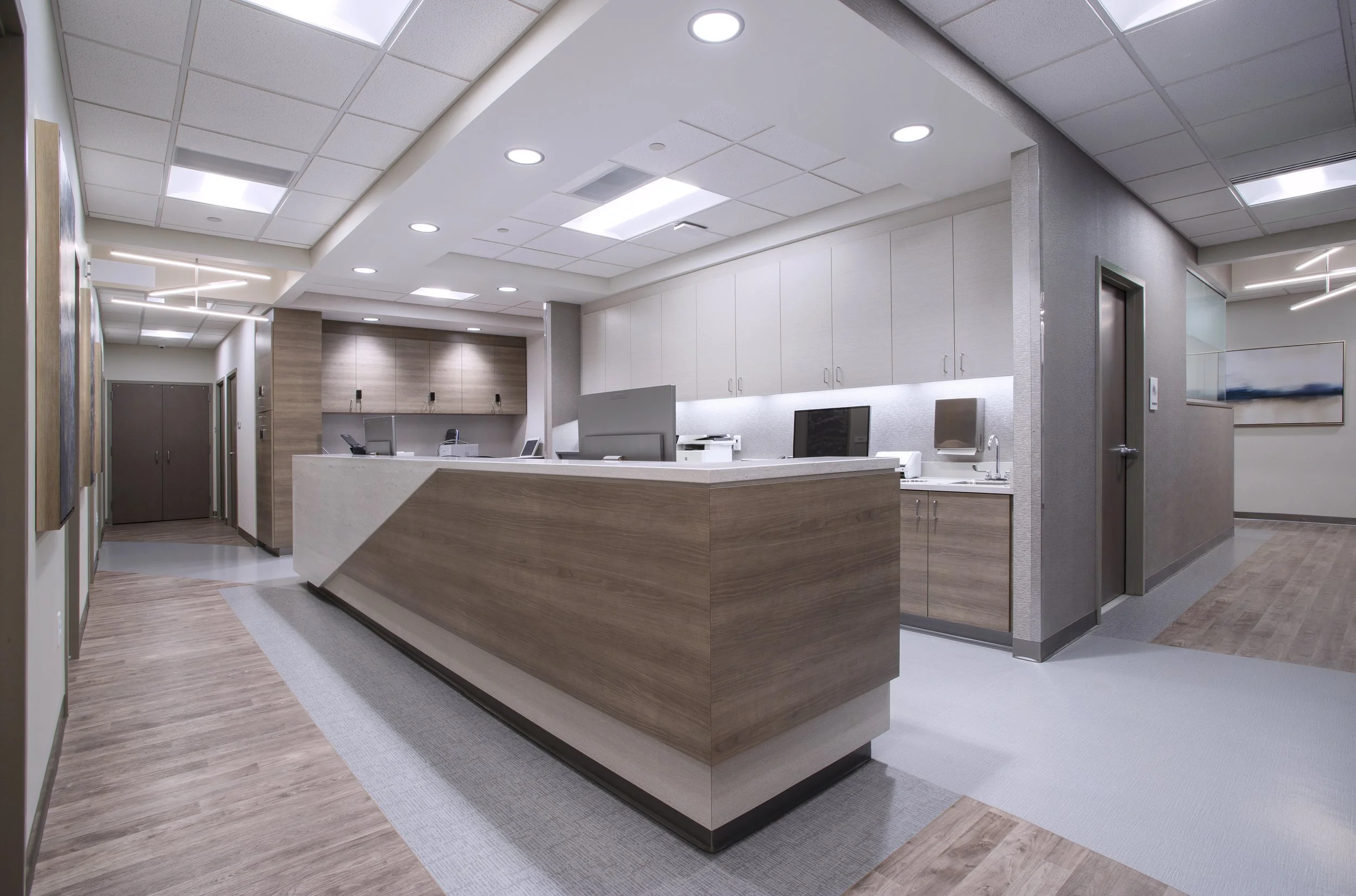 Modern hospital reception area with a large wooden counter, computer monitors, and white cabinets, with artwork hanging on the walls and a wood and tile floor.