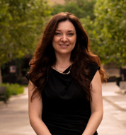Woman with long brown hair wearing a black sleeveless top standing outdoors in a park with trees and pathway in the background.