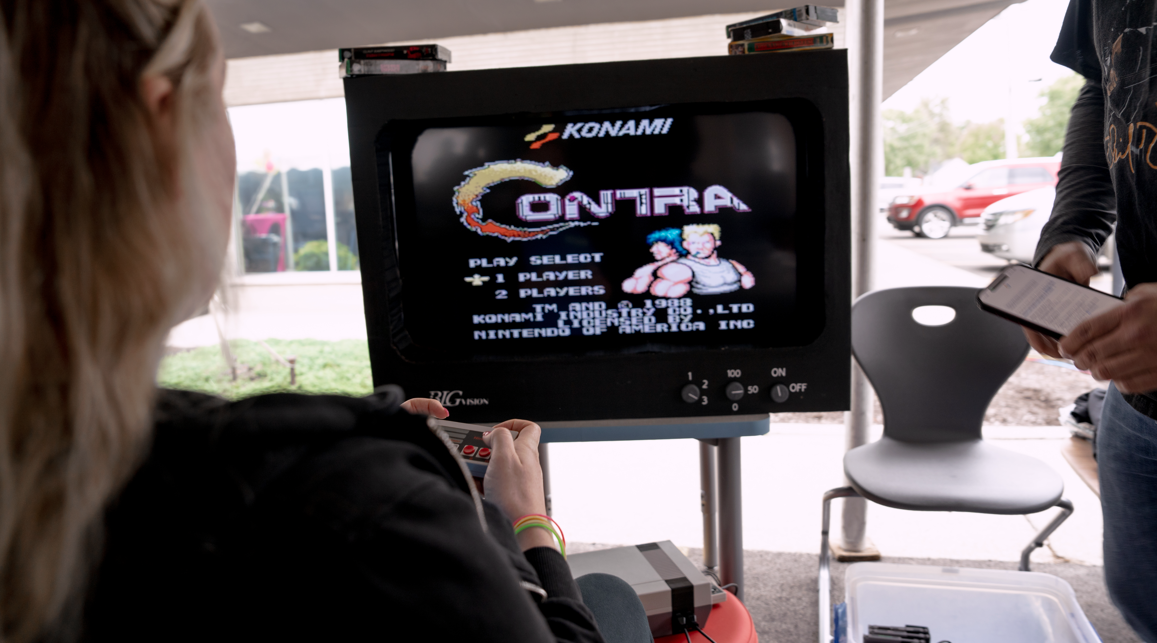 Person playing Konami Contra video game on a CRT TV while another person looks at a phone outside.