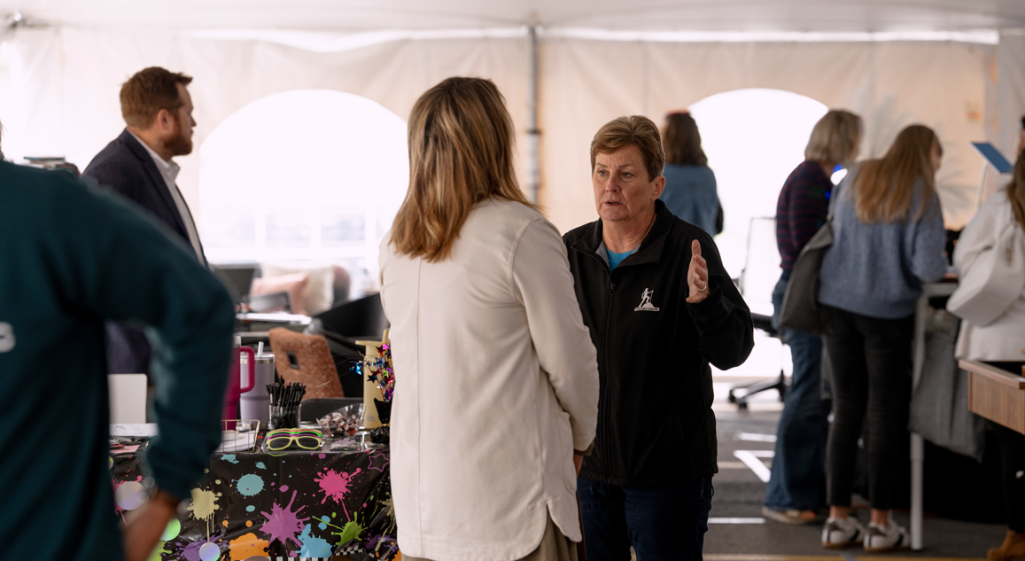 Two women are engaged in conversation inside a large tent, with several people and tables in the background.