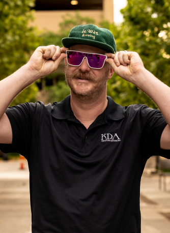 Man wearing a green beanie, purple sunglasses, and a black polo shirt with a logo, adjusting his sunglasses outdoors.