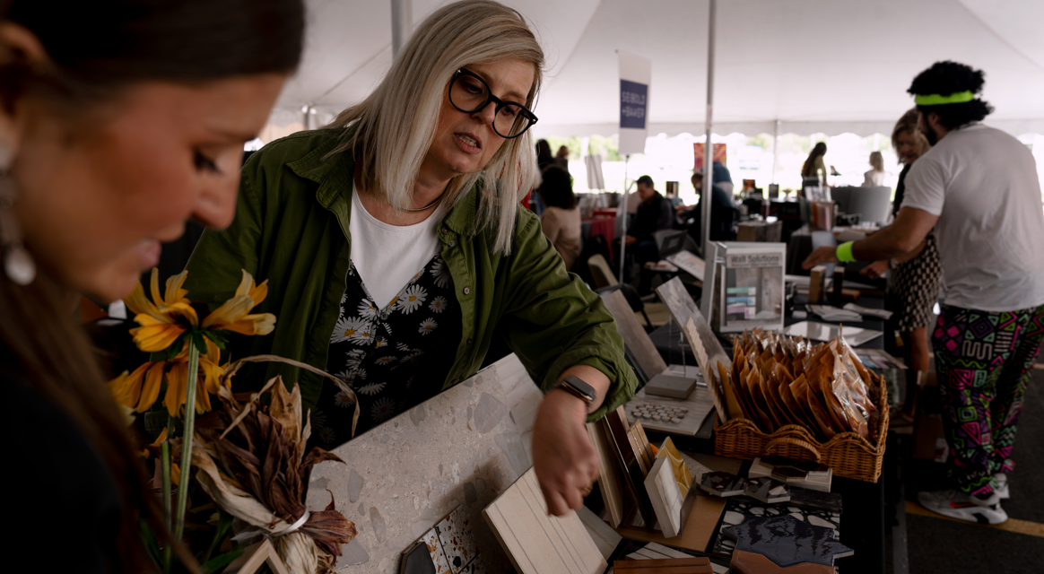 A woman with gray hair, glasses, and a green jacket is interacting with a vendor at a table that has picture frames and a basket of packaged items, while a person dressed in colorful, eccentric clothing stands in the background in a busy outdoor mark