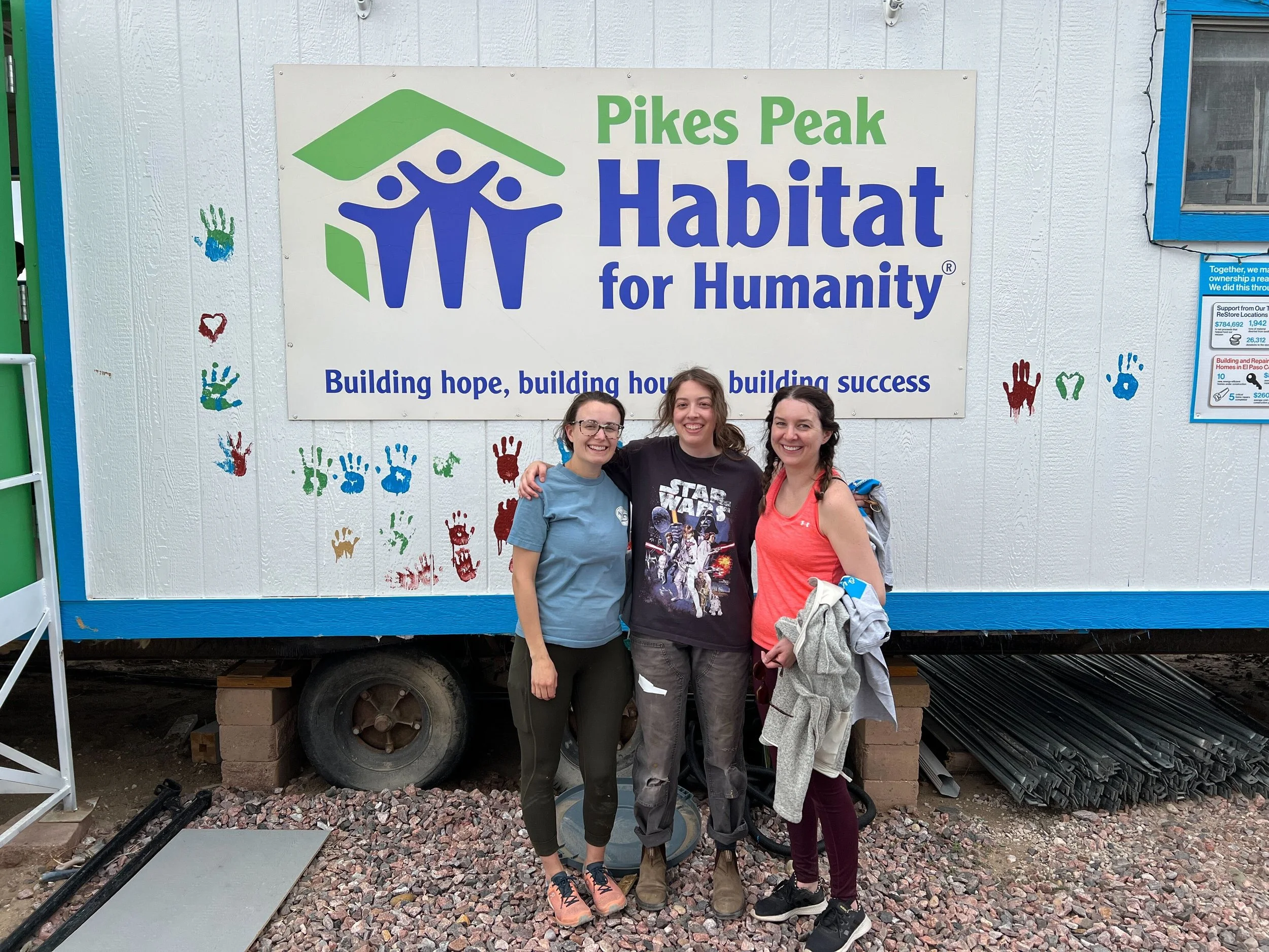 Three women standing together outdoors in front of a large sign that reads 'Pikes Peak Habitat for Humanity,' with colorful handprints painted on the white wall behind them. They are smiling and appear happy.
