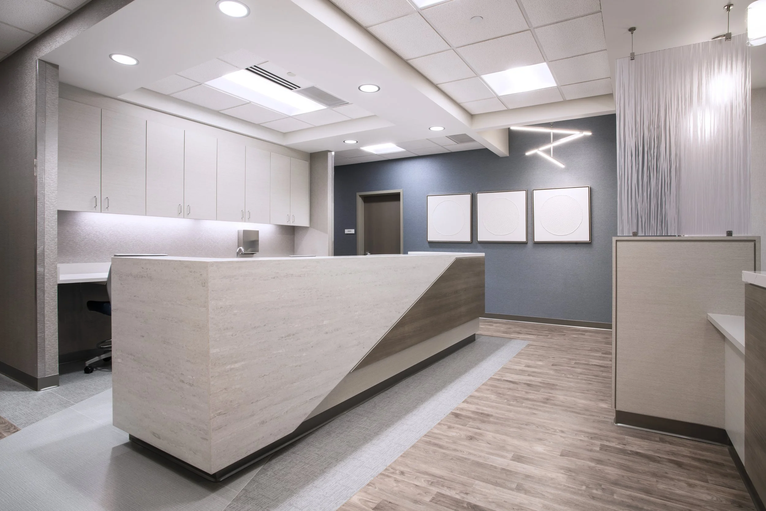 Modern medical or office reception area with white cabinets, a curved reception desk, gray walls, framed abstract artwork, wooden flooring, and contemporary lighting fixtures.