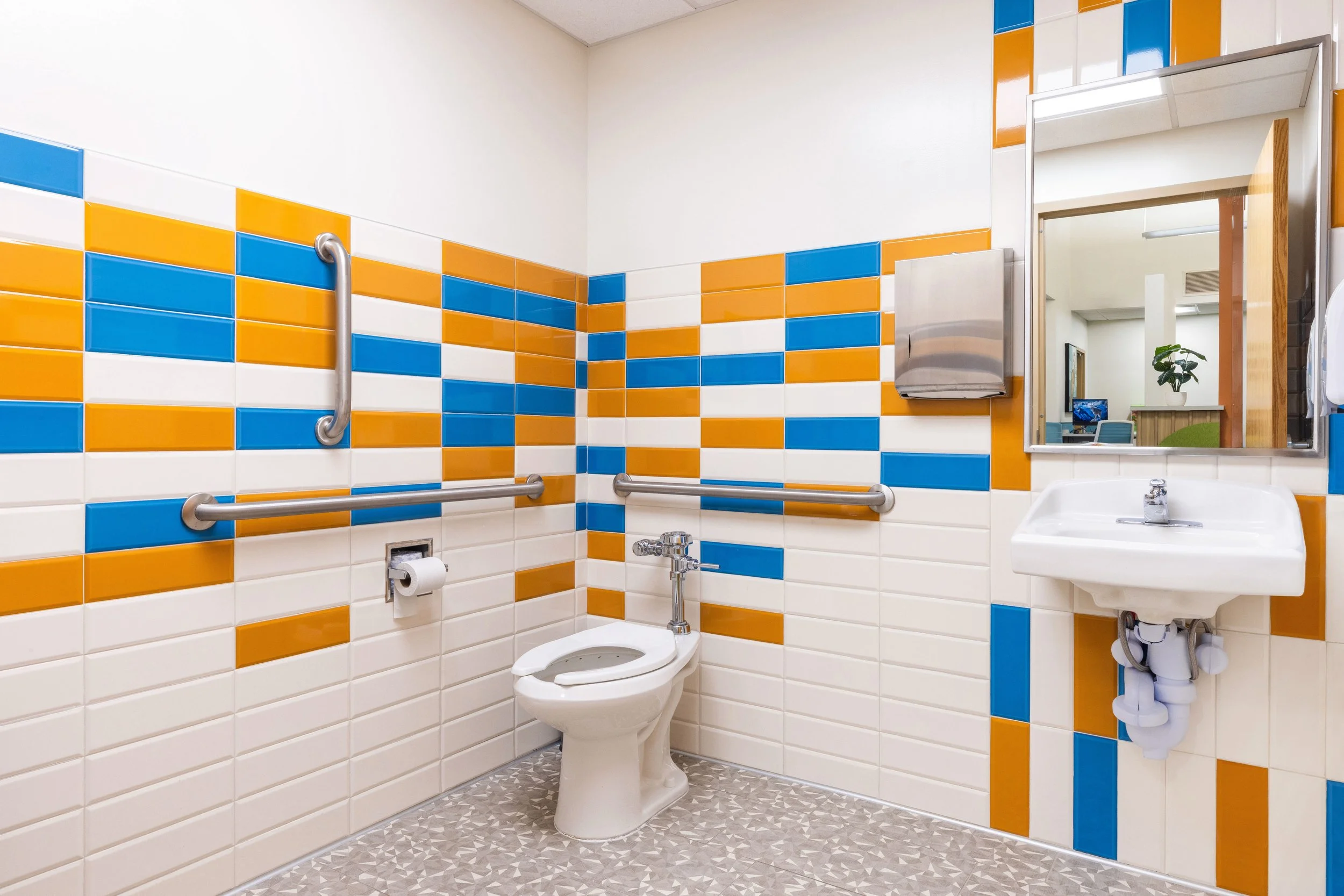 A brightly colored accessible restroom with white, orange, and blue tiles on the walls, equipped with grab bars, a mirror, a toilet, a small sink, and a paper towel dispenser.