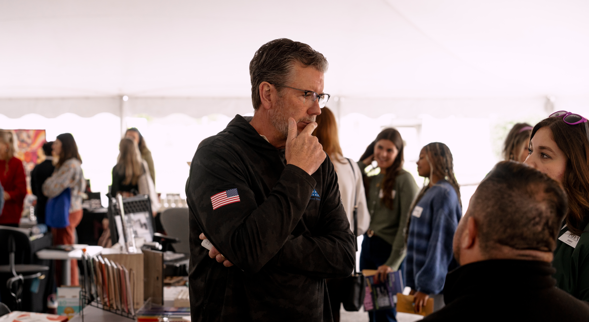 A man with glasses, gray hair, and a beard, wearing a black jacket with an American flag patch on the sleeve, stands in a crowded indoor event tent, thinking or contemplating with his hand on his chin, surrounded by women and people engaging in conve