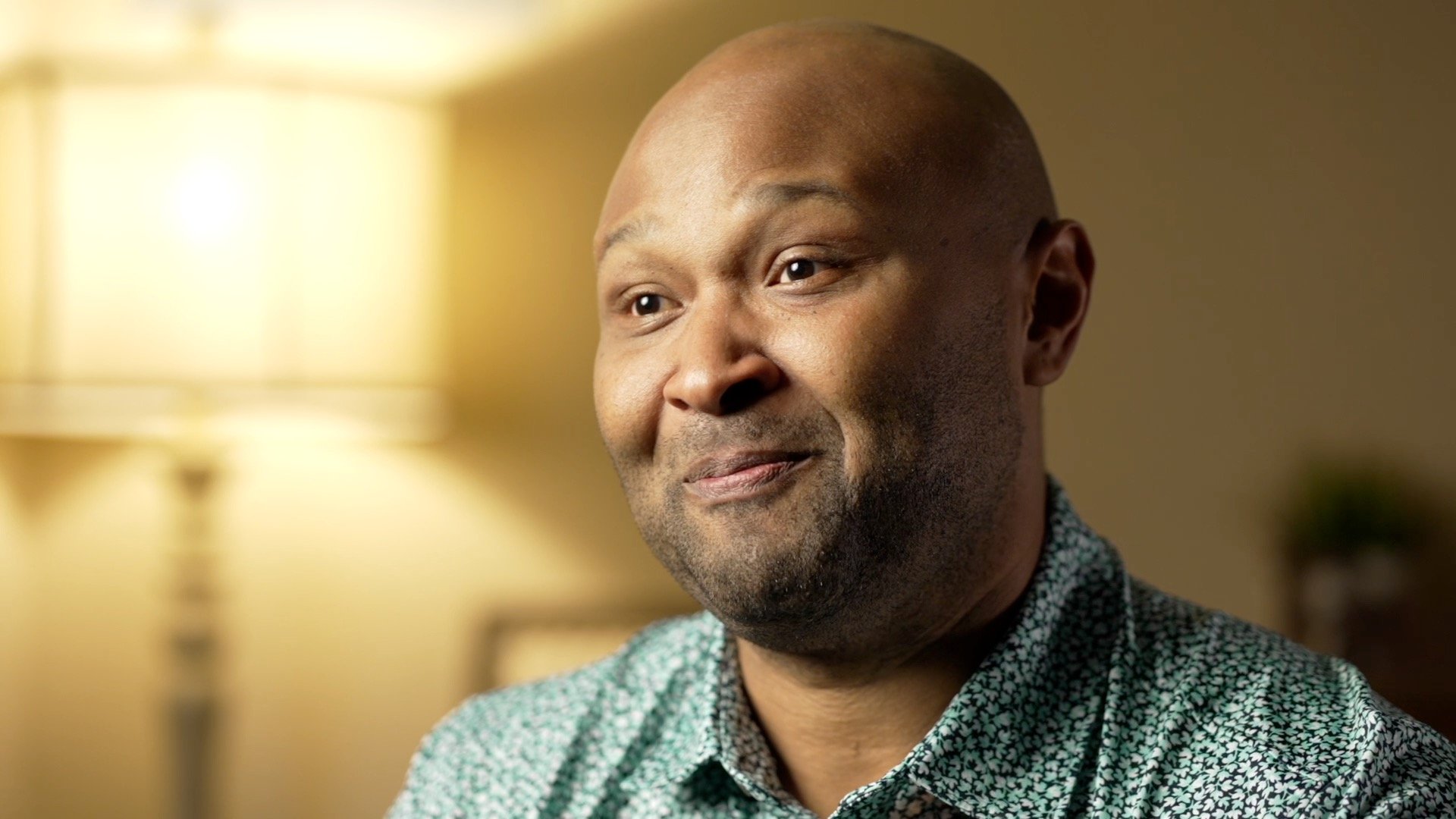A close-up of a smiling African American man with a bald head, wearing a patterned shirt, in a warmly lit indoor setting.