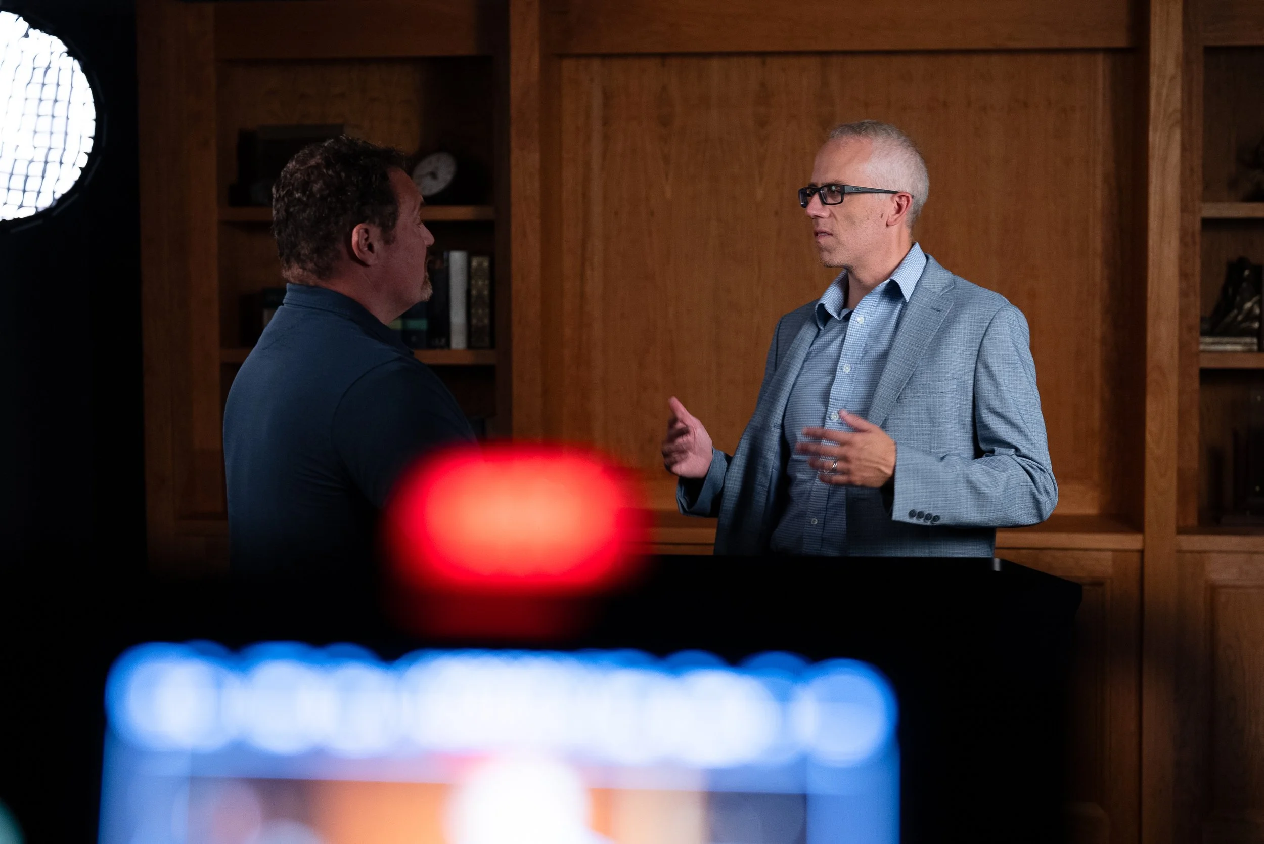 Two men are having a serious conversation in a wood-paneled room. One man is wearing glasses and a light blue blazer, while the other has curly hair and is dressed in dark clothing. A soft light illuminates their faces, and bookshelves are visible in the background. The photo appears to be taken through a camera lens, with colorful bokeh effects at the bottom.
