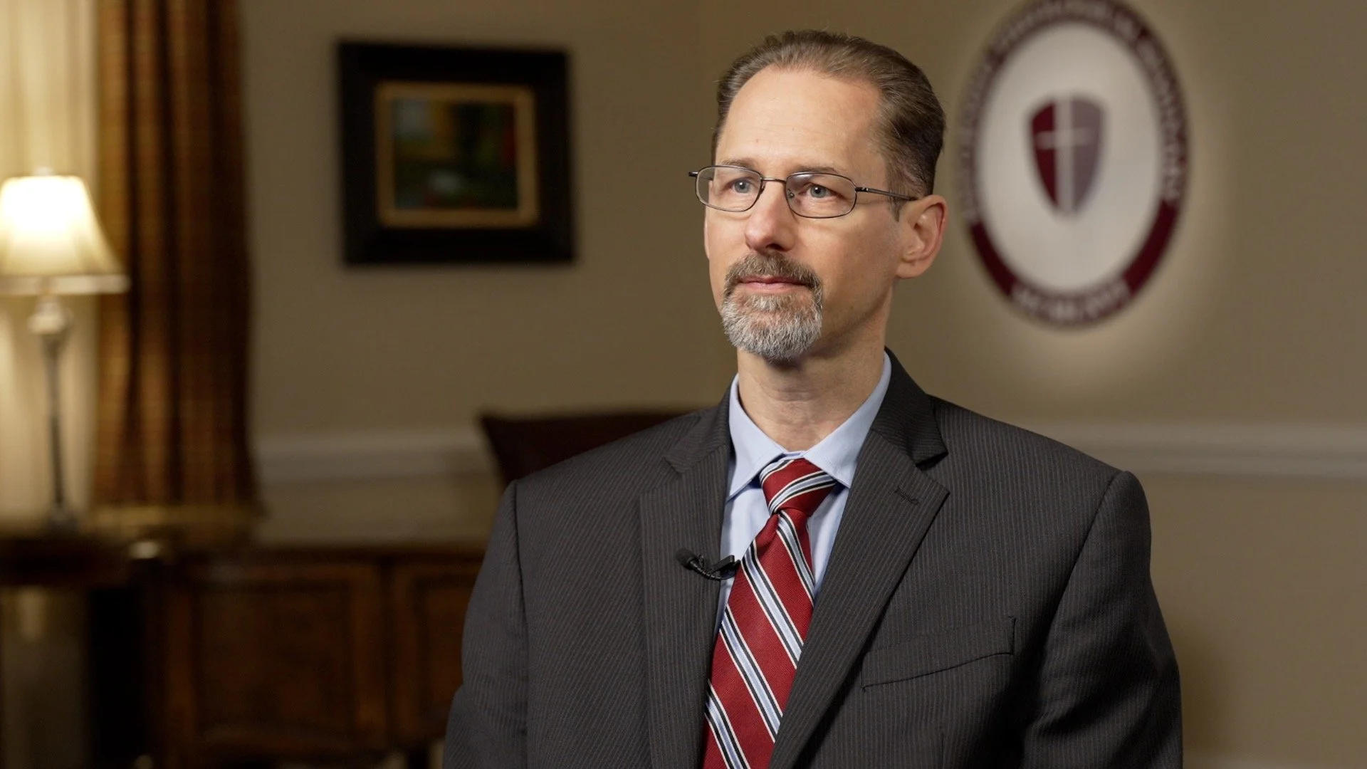 A man in a gray suit with a red and white striped tie, glasses, and a beard, sitting in a formal room with a crest on the wall behind him.