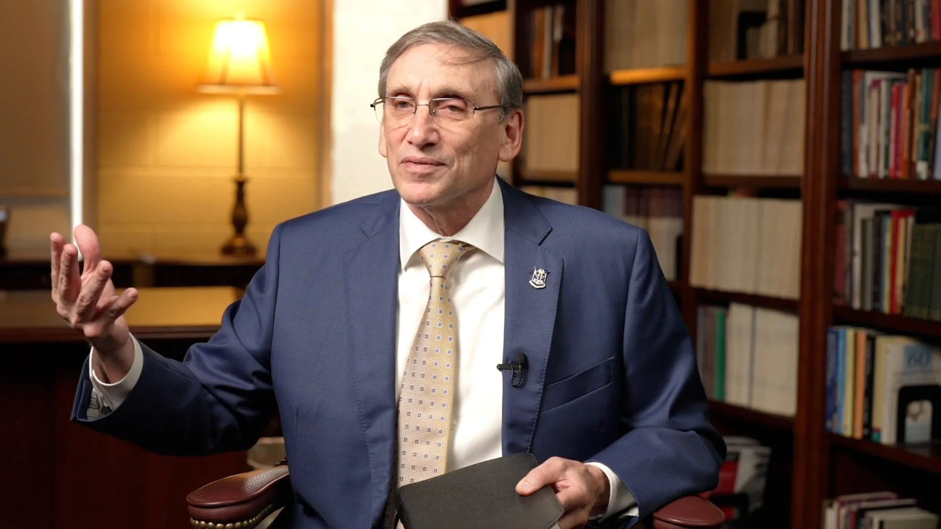 A man in a blue suit, white shirt, and patterned tie, with glasses, is speaking and gesturing with one hand while seated in an office or library setting with bookshelves and a lit lamp in the background.