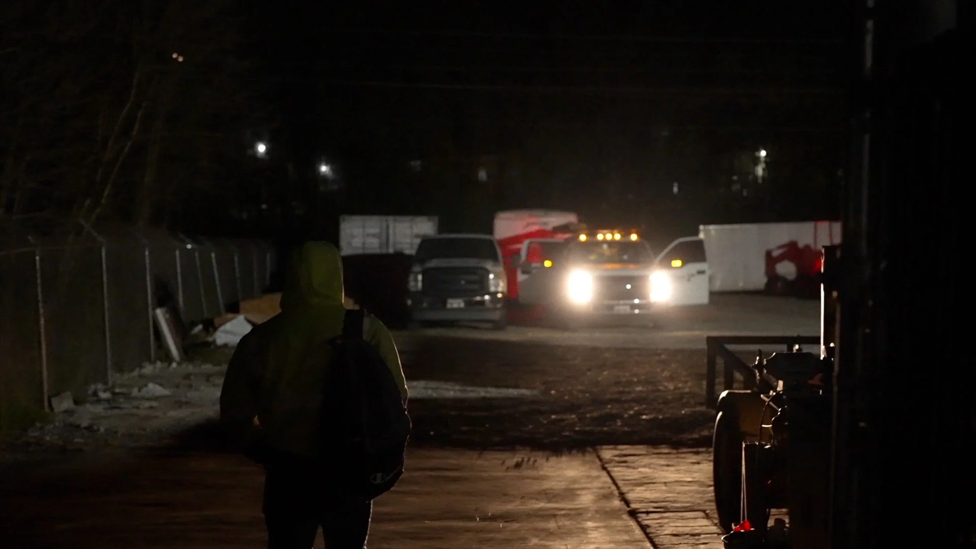 A person walking on a dark street at night, with a row of trucks and trailers visible in the distance, illuminated by headlights.