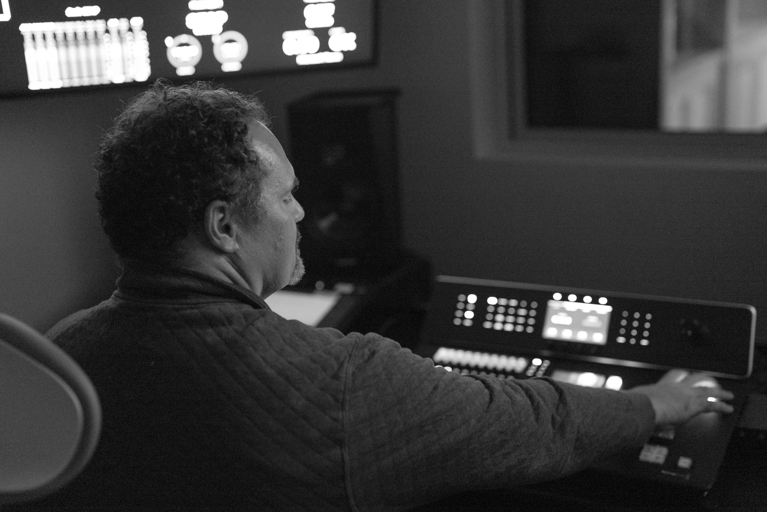 A man with curly hair working at a control panel in a dark room, with monitor screens displaying technical data.