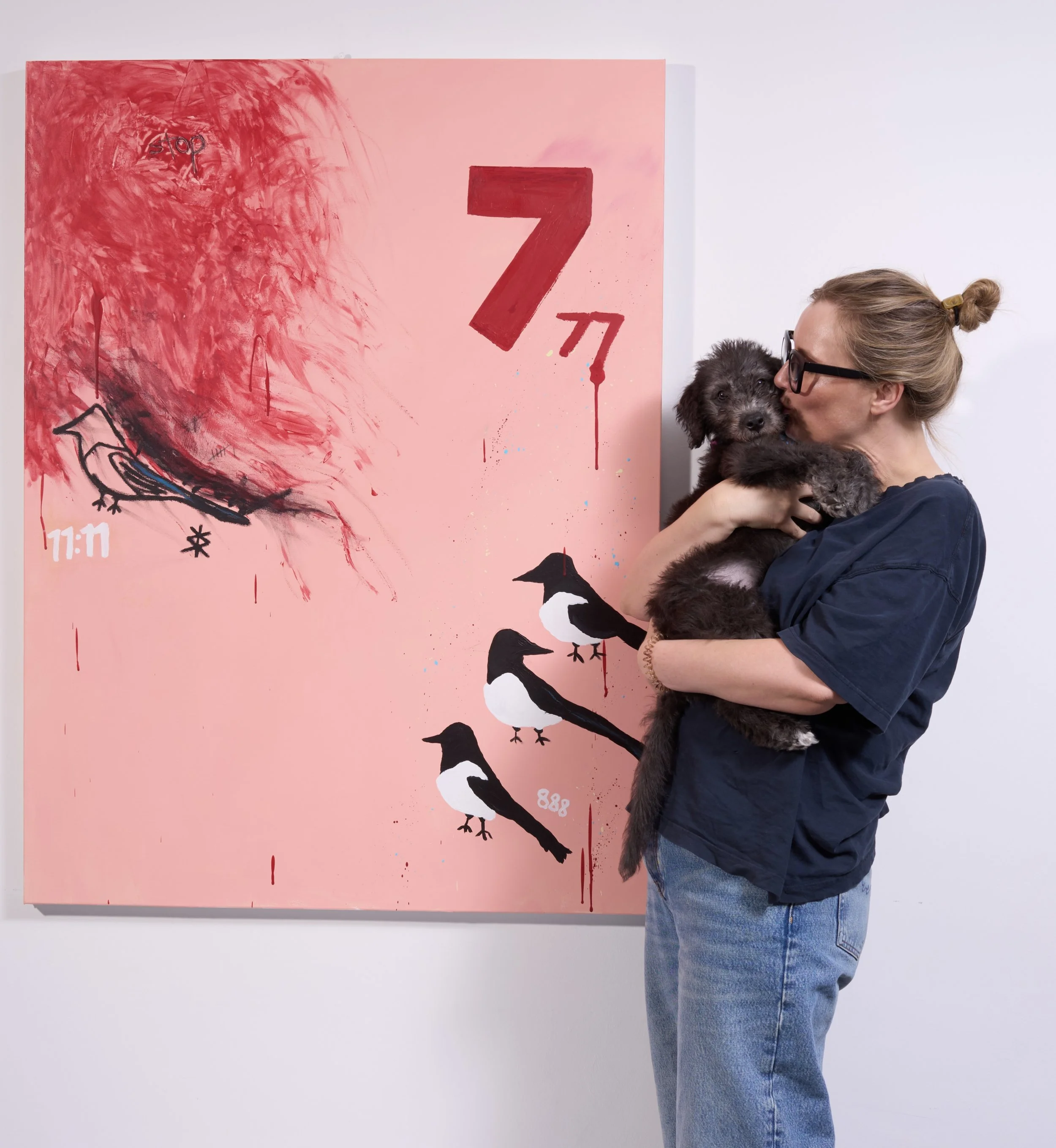 Artist in front of a pink painting with abstract brush strokes and graffiti-style elements, featuring magpies, a large red number 7, and various small symbols.