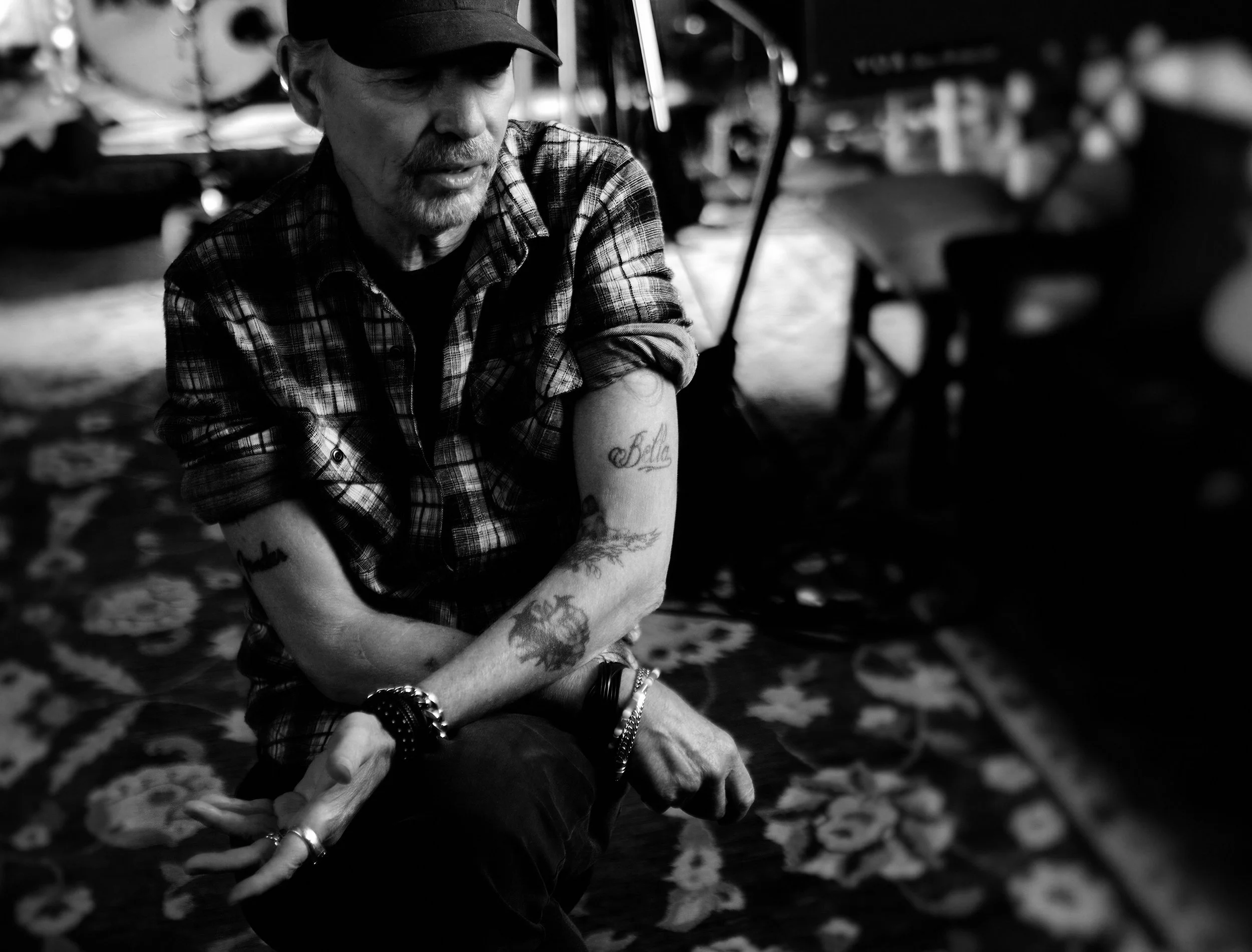 A man with tattoos on his arms, wearing a plaid shirt and a cap, sits with his arms crossed in a black and white photograph. He appears to be in a contemplative mood, surrounded by musical equipment.