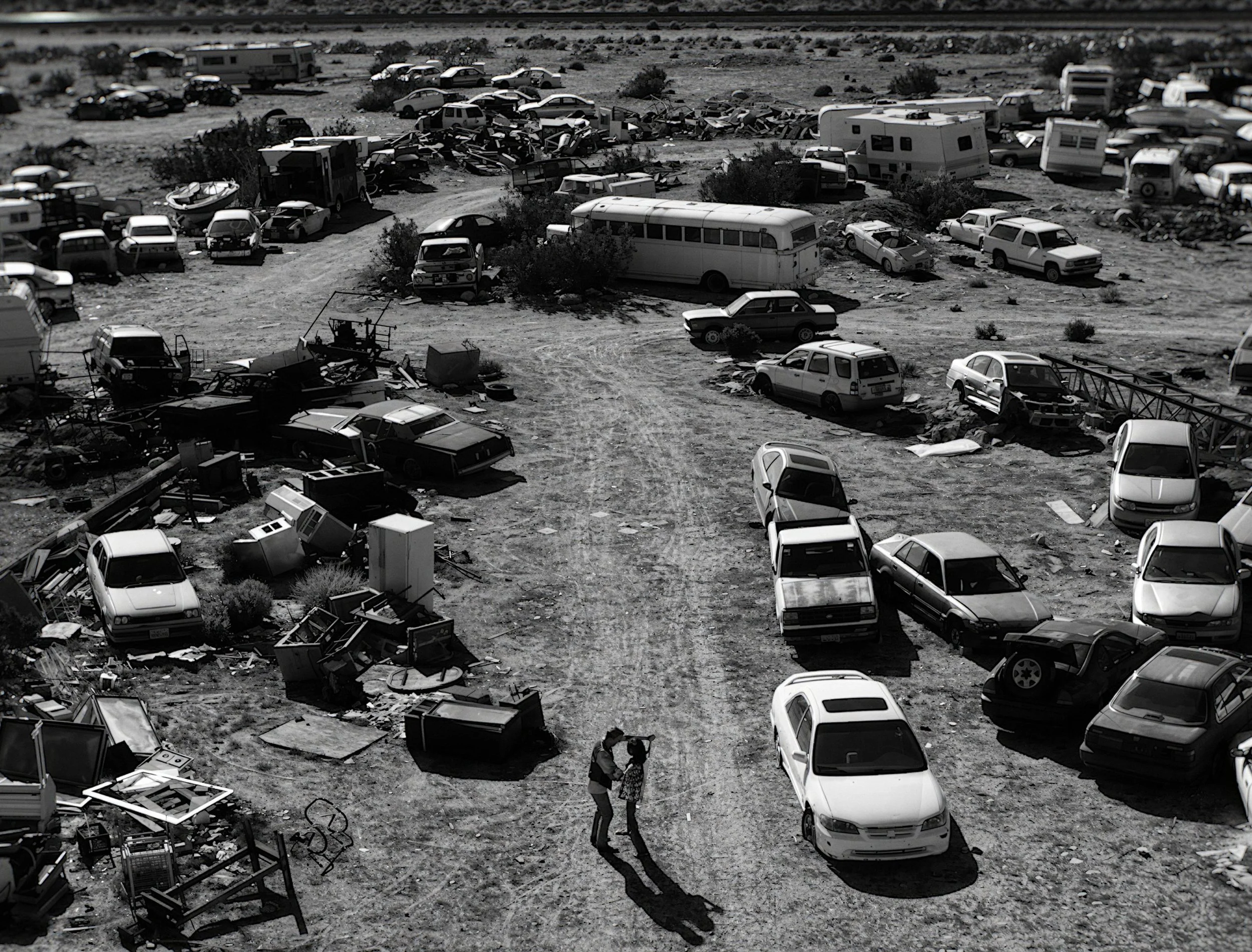 A desolate desert scene with numerous abandoned, wrecked, and damaged cars, trucks, and RVs scattered across the ground. Some vehicles are tipped over or broken down, with debris and discarded objects lying around, suggesting a junkyard or crash site