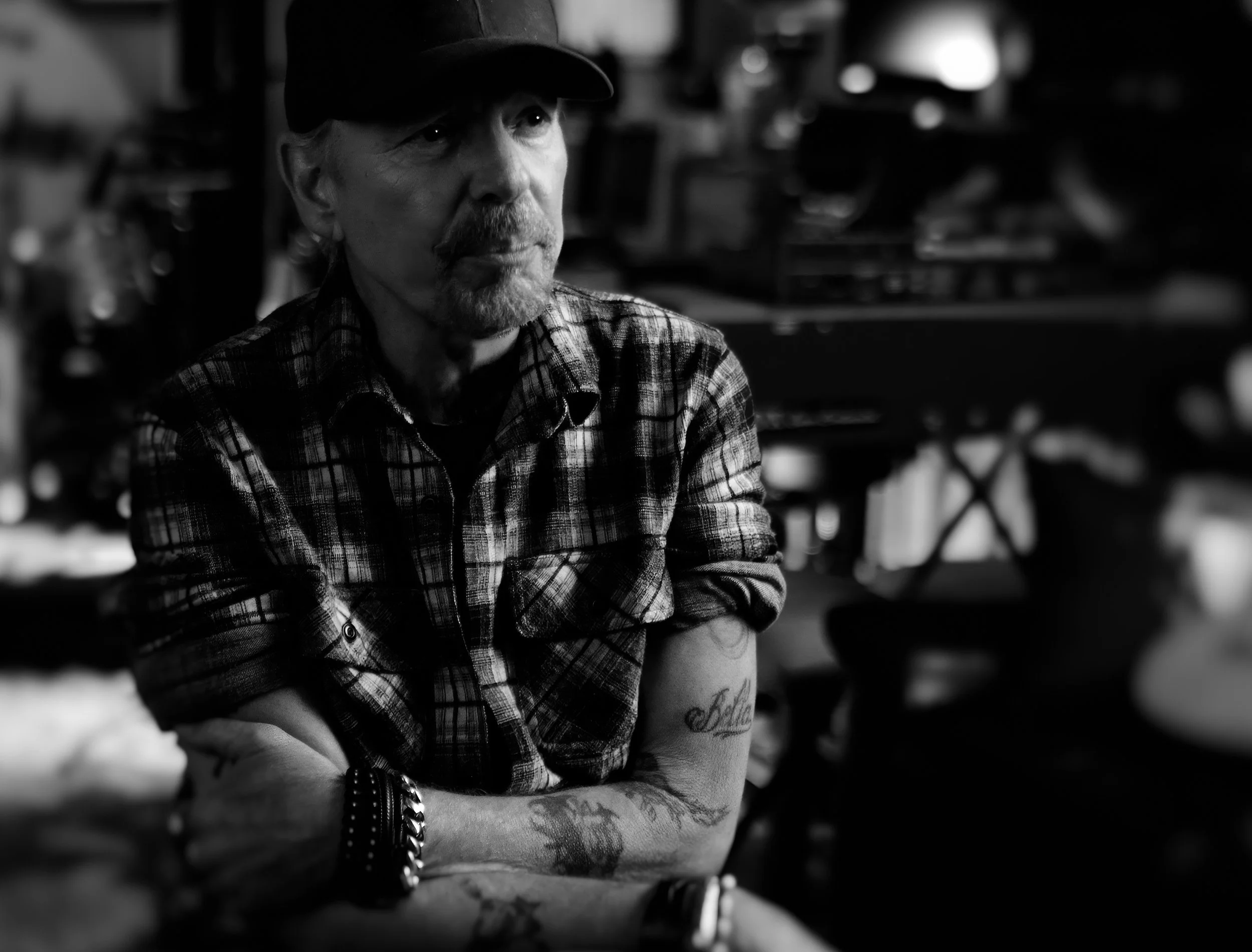 A black and white photo of a man with a beard and mustache wearing a cap and plaid shirt. He is sitting with arms crossed, showing tattoos on his forearms, and looking off to the side. The background is blurry with shelves and objects.