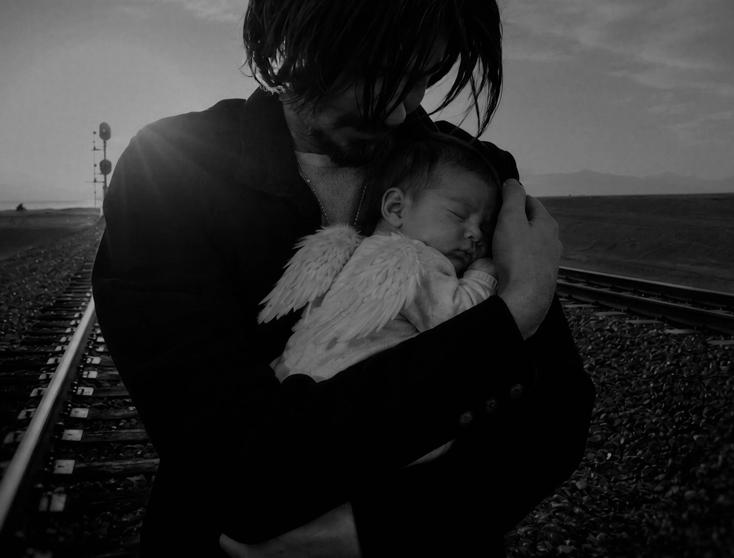 A person with long dark hair is holding a sleeping child with angel wings, standing on railroad tracks outdoors during sunset or sunrise.