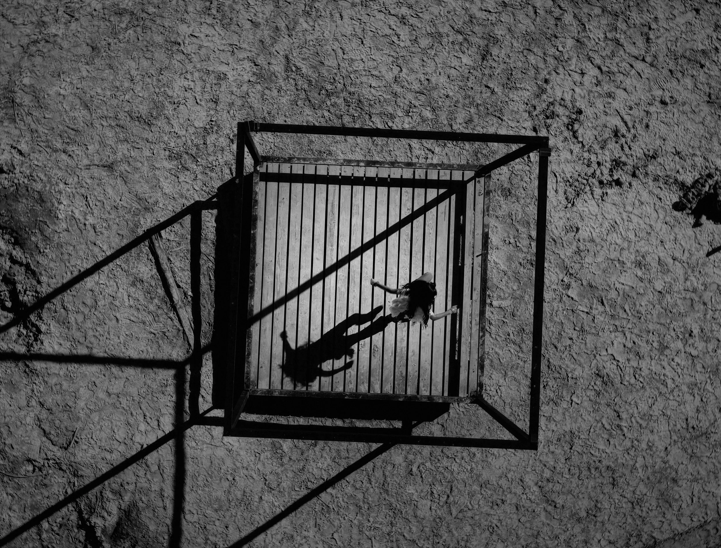 A person lying on their back inside a playground structure made of metal bars, viewed from above on a dirt surface.