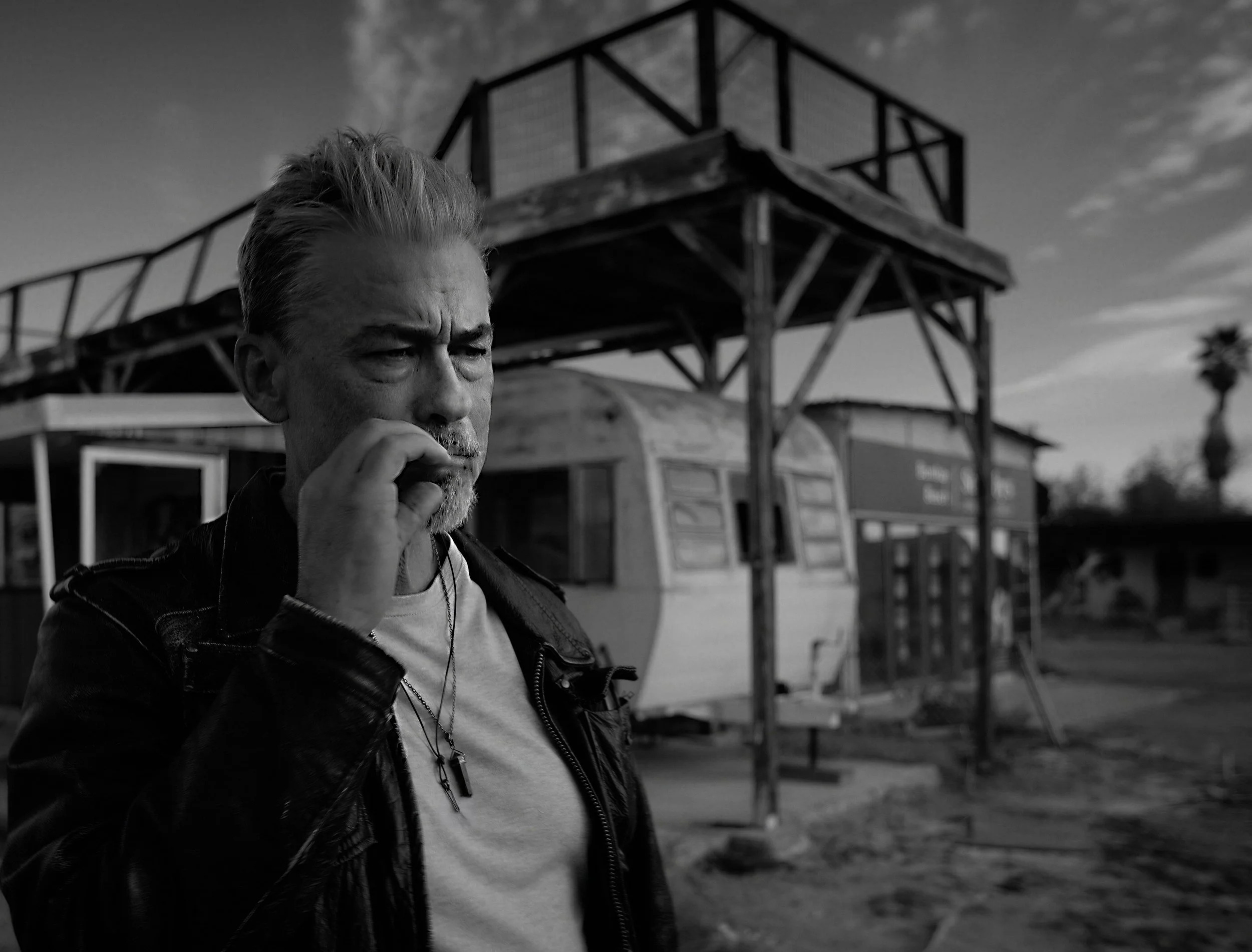 A black and white photo of a man with gray hair and a beard, holding his hand near his face, standing outdoors near a wooden structure with a staircase and railing.
