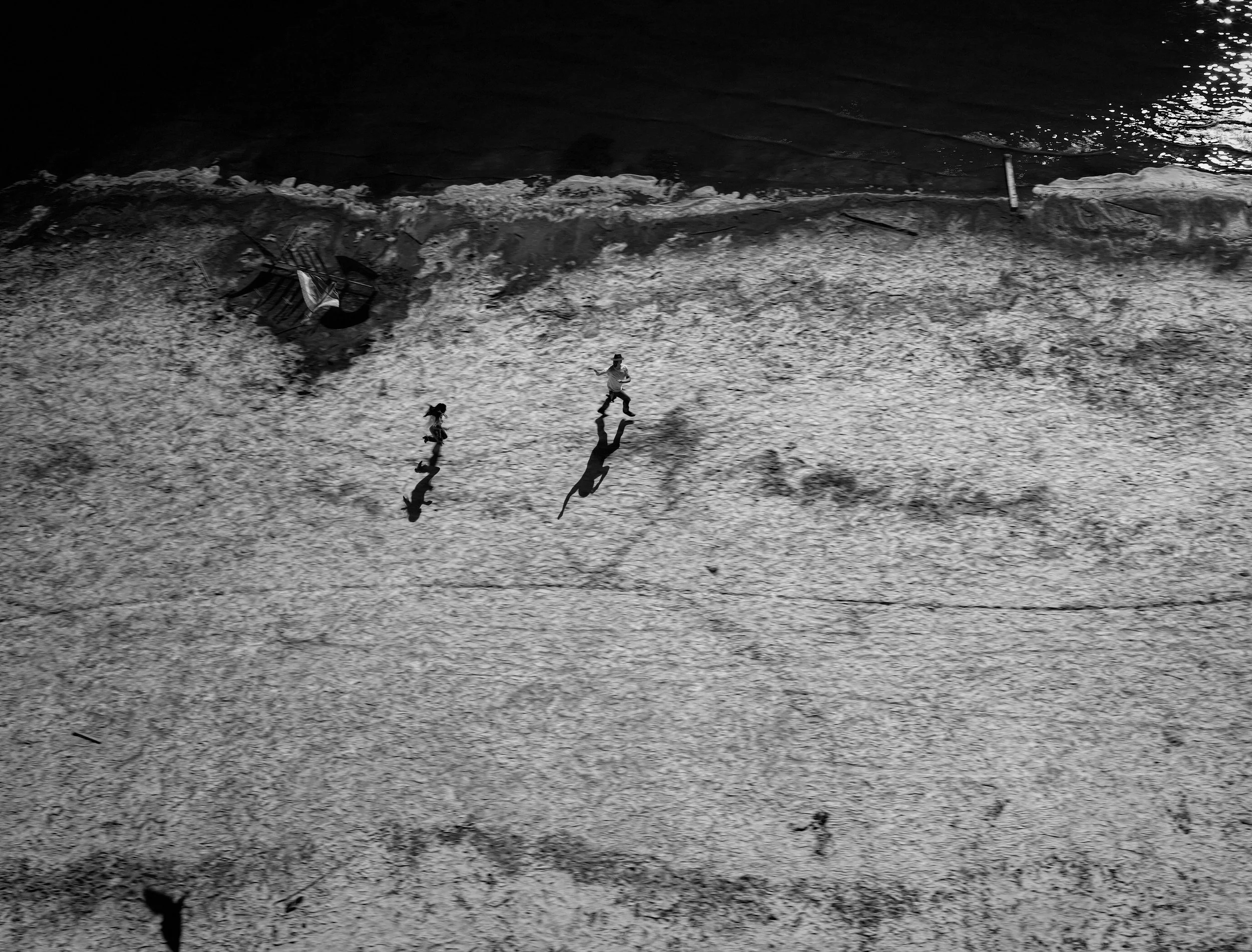 Black and white photograph of a beach with two children running and playing, with their reflections visible on the wet sand.
