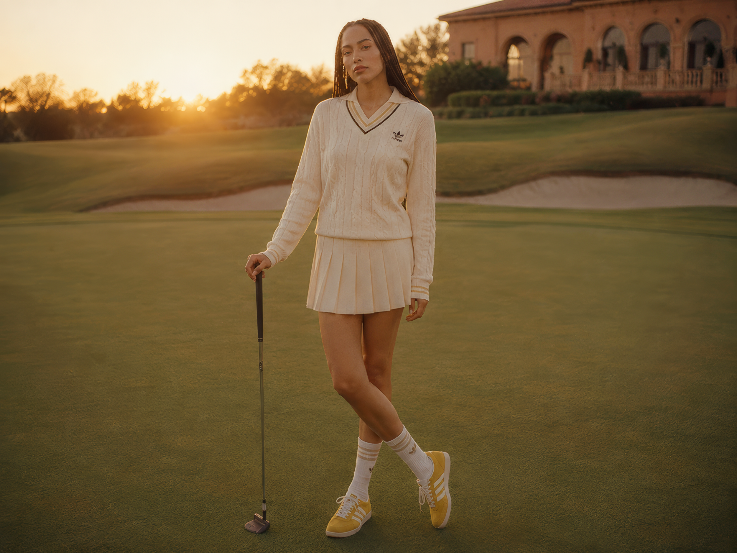 A young woman with long braided hair wearing a cream-colored golf sweater and skirt, standing on a golf course at sunset, holding a golf club, with a large house in the background.