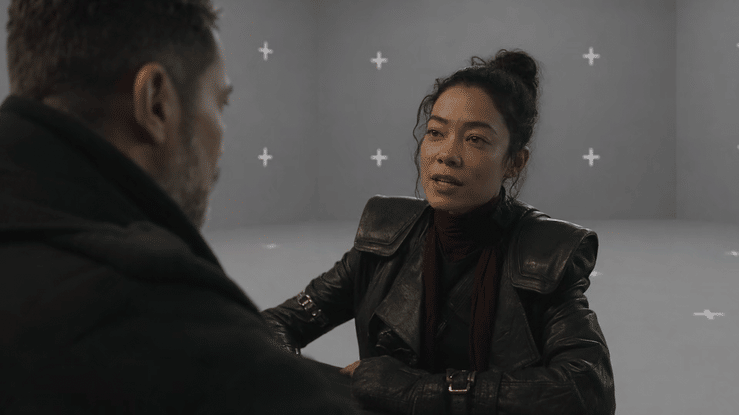 A woman and a man having a conversation in a gray room with plus signs on the walls.