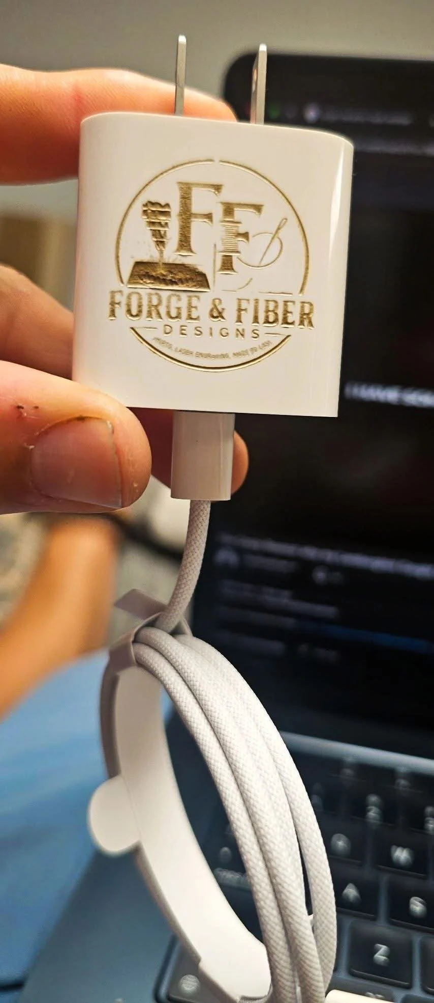 A person holding a white power adapter with the logo 'Forge & Fiber Designs' and a plug, with white earphones hanging from it, inside a vehicle.