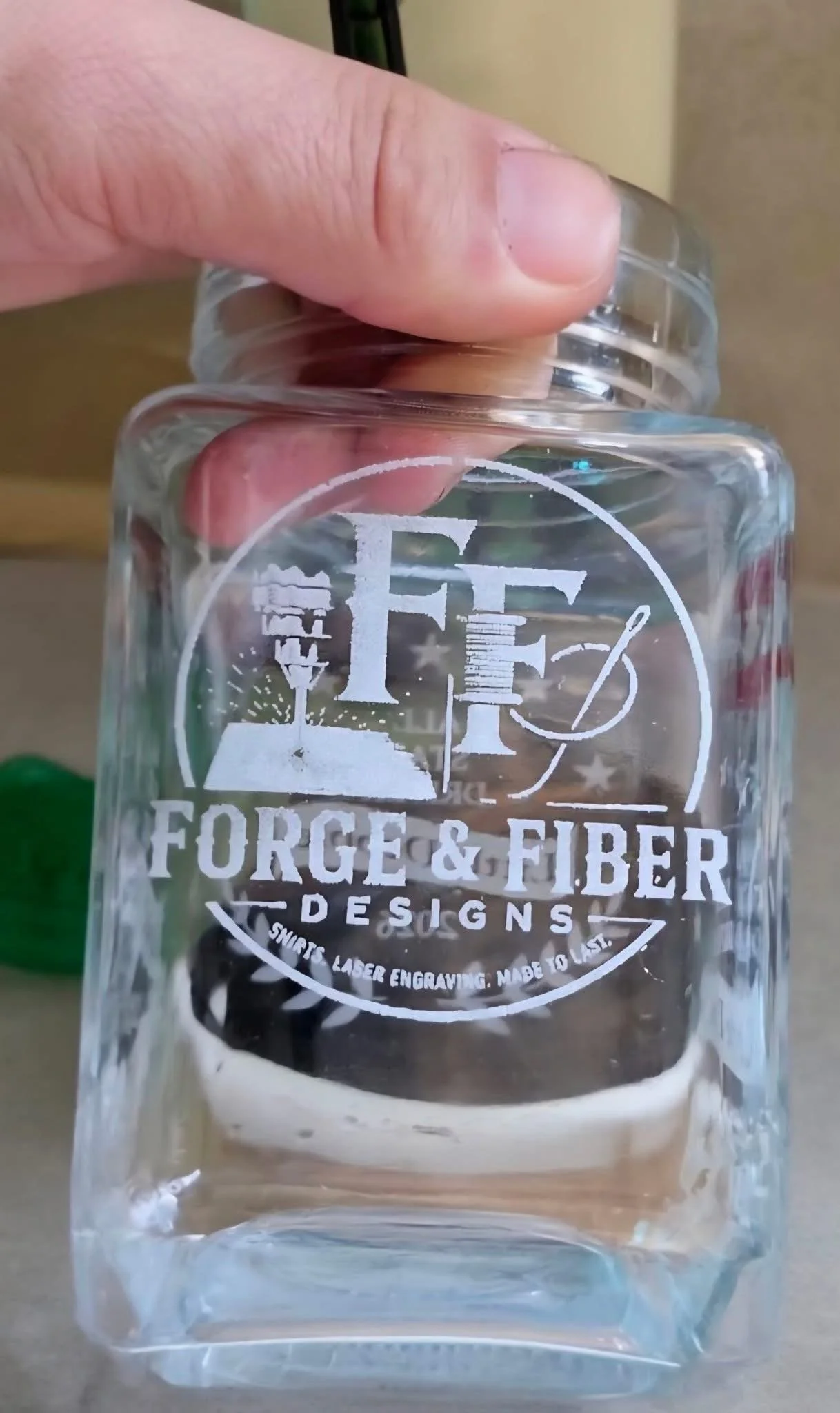 Close-up of a clear glass mug with white logo text from Forge & Fiber Designs, showing the person's thumb and fingers holding it against a background of a tan wall and a computer monitor.