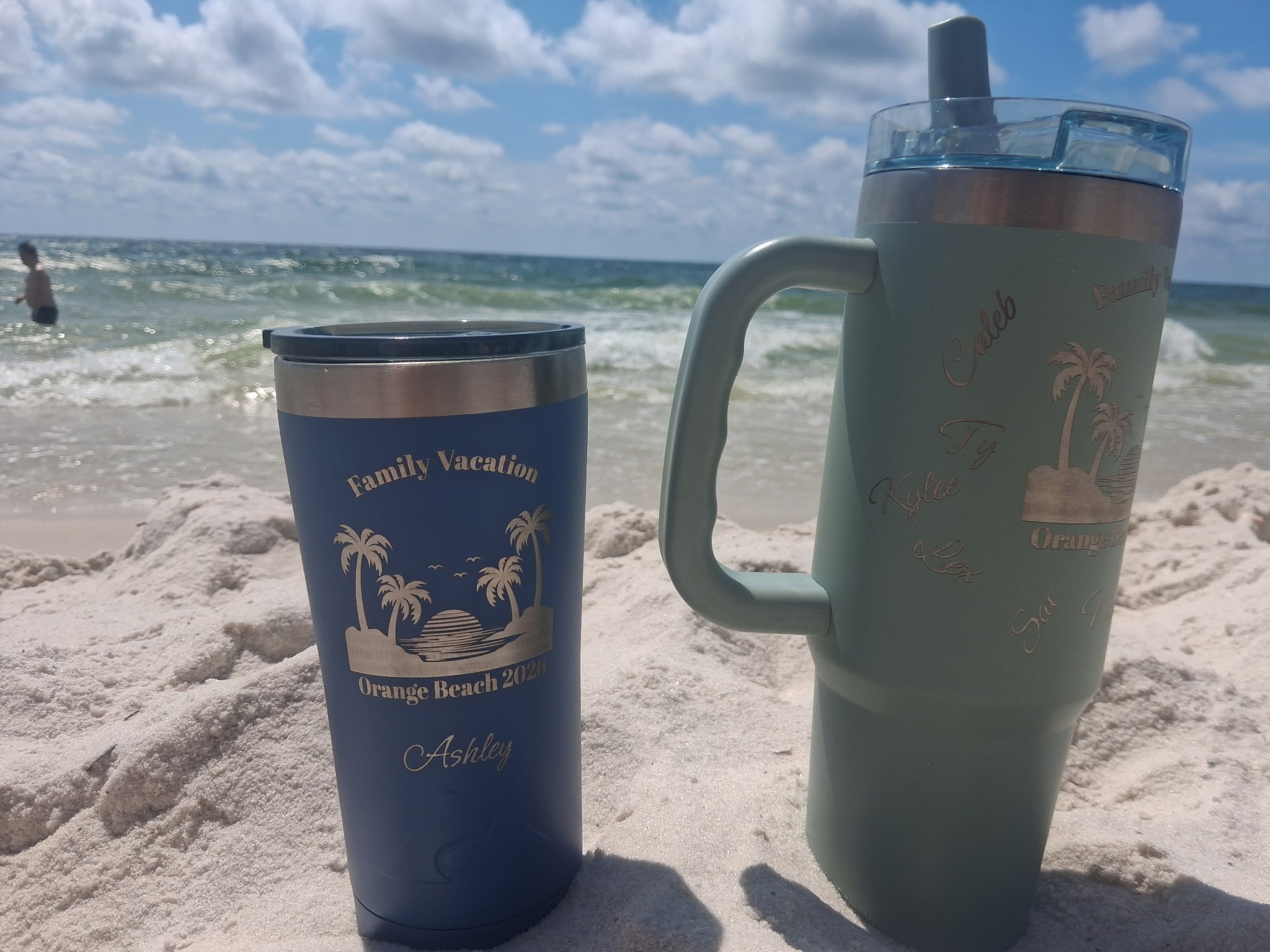 Two travel mugs with personalized text and palm tree illustrations resting on the sand at a beach, with the ocean, a person in the water, and a partly cloudy sky in the background.