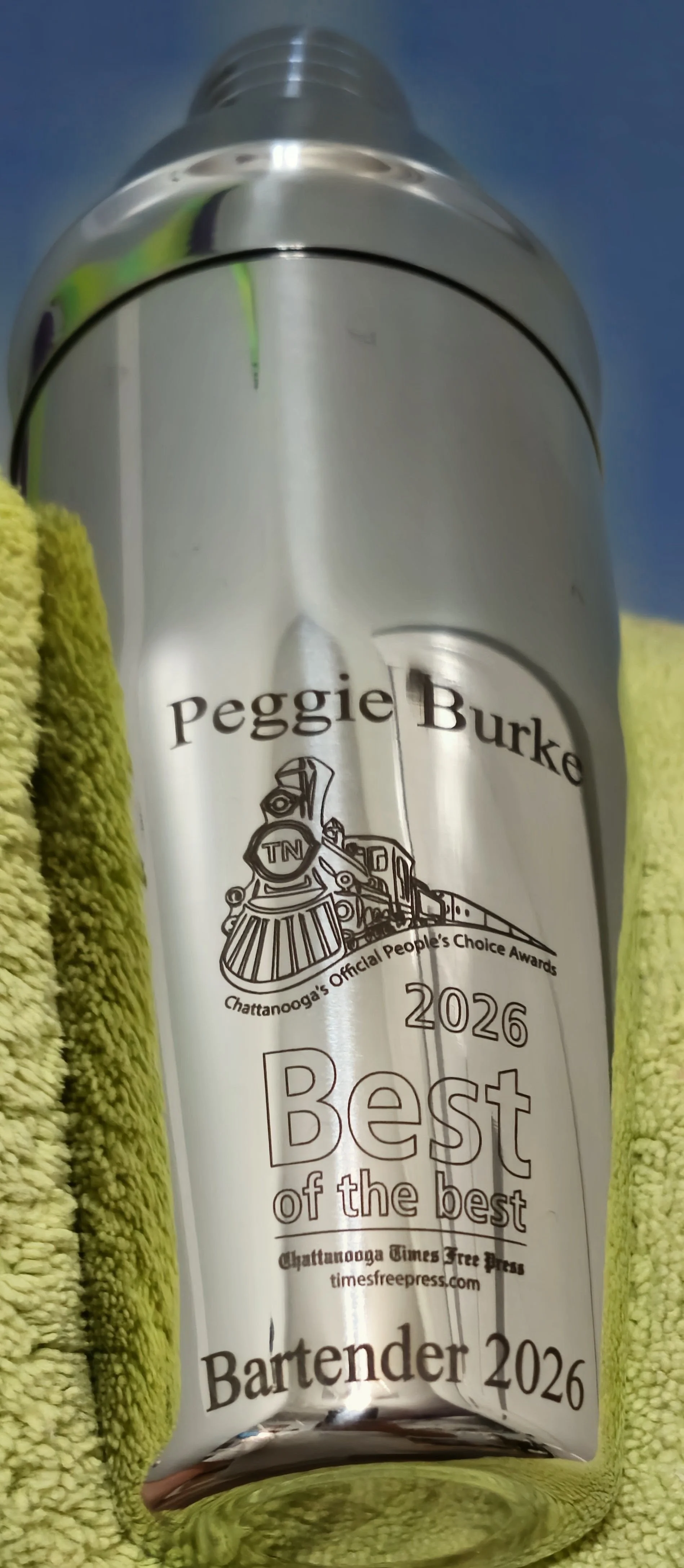 A metallic can with a train illustration and the text 'Peggie Burke 2026 Best of the best Bartender 2026' printed on it, held by yellow towels.