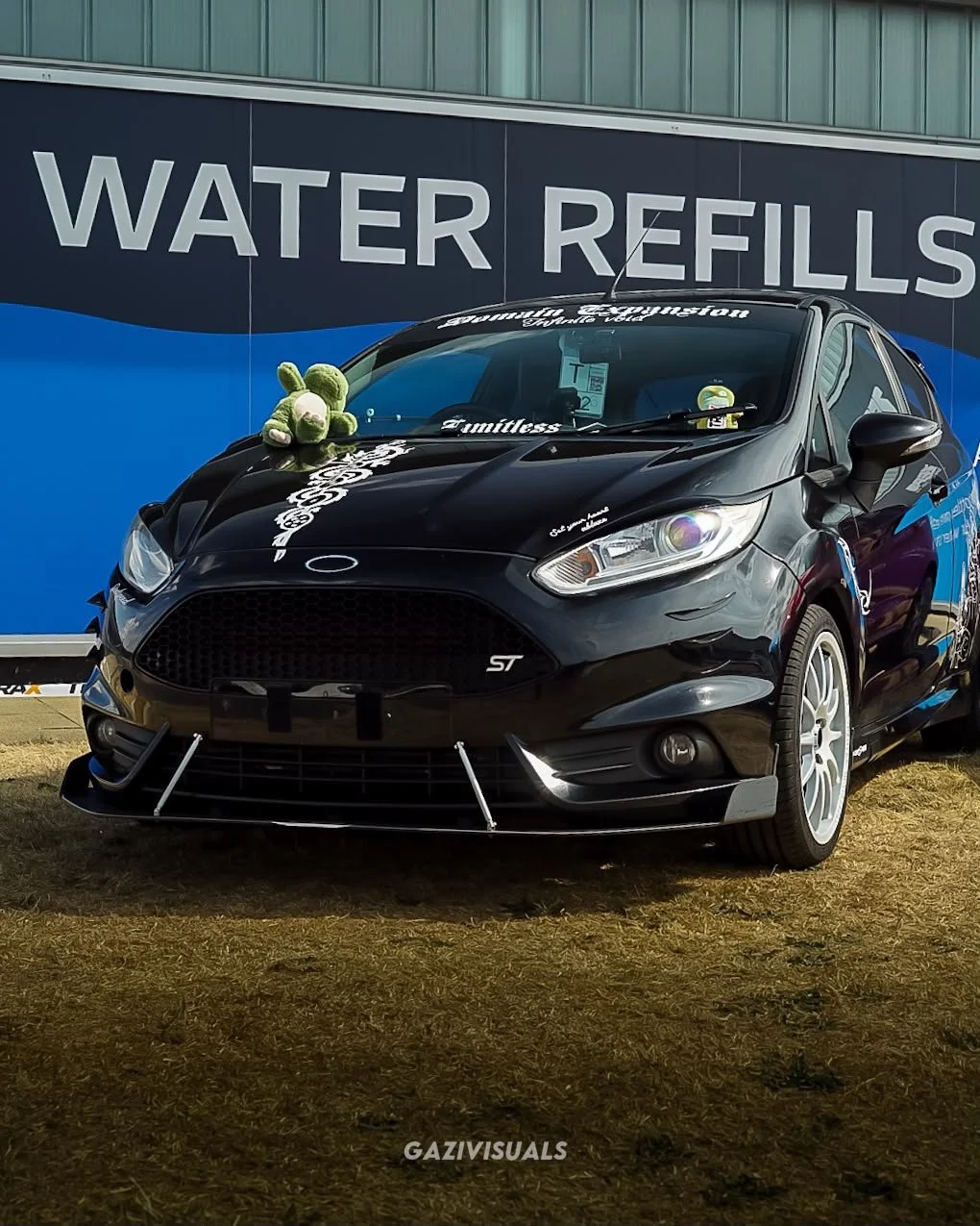 A black Ford Fiesta ST with a plush toy on the hood, parked in front of a sign that reads "Water Refills."