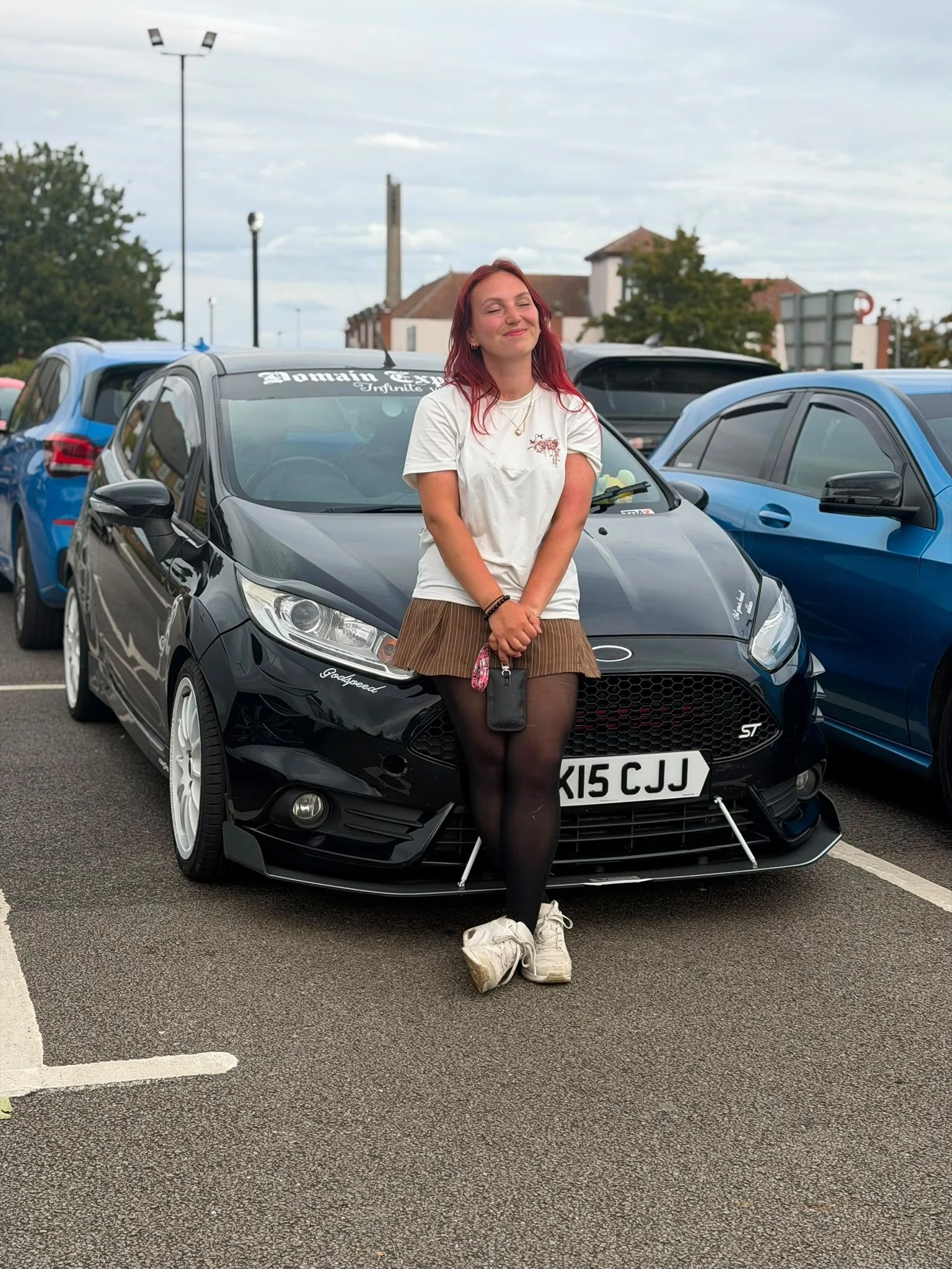 A young woman with red hair, wearing a white t-shirt, brown skirt, black tights, and white sneakers, standing in a parking lot in front of a black car. She has a happy expression and is holding a pouch with her shoes tied to her bag.