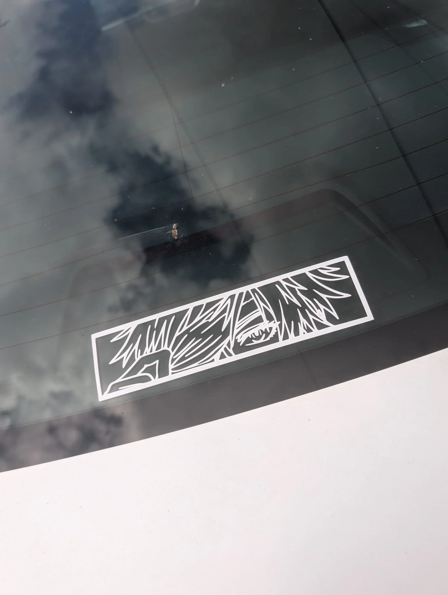 A white sticker with black graffiti-style text that says 'RAVEN' on the rear window of a car, with reflections of the sky and clouds.