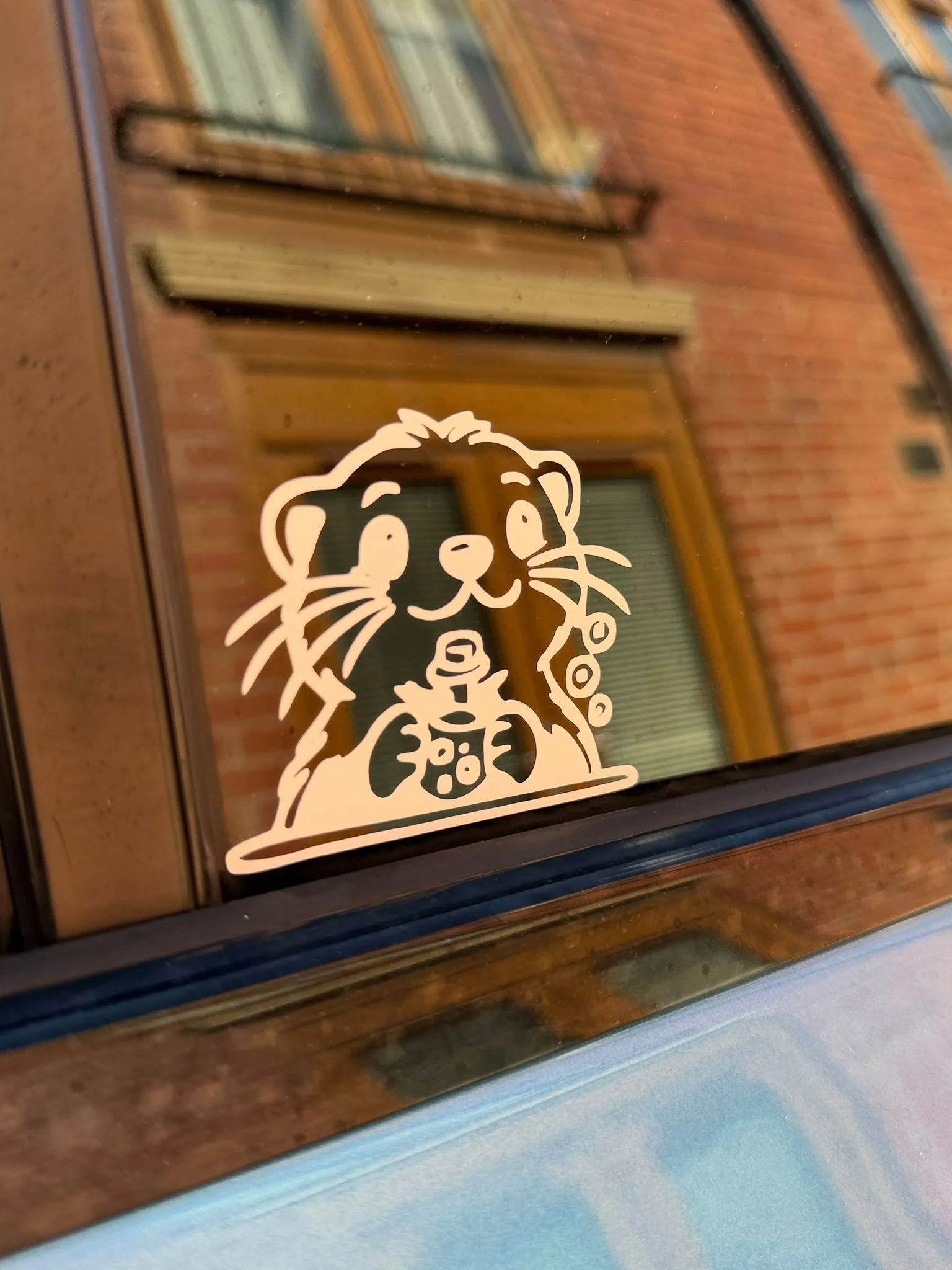 A sticker of a cartoon animal, possibly a beaver, holding a mug, on a reflective glass surface with a brick building featuring orange and blue window frames in the background.