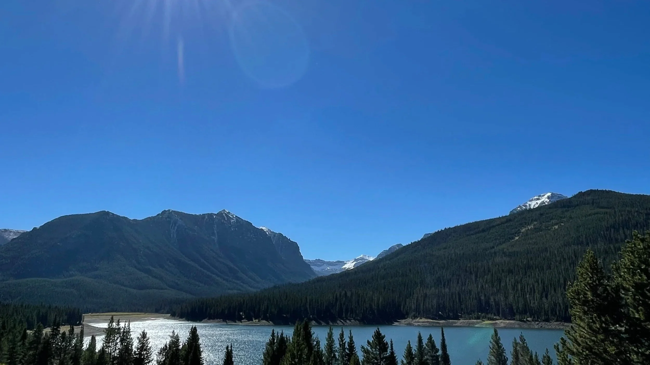 Mountain landscape with snow-capped peaks, forested slopes, and a lake in the foreground under a clear blue sky.