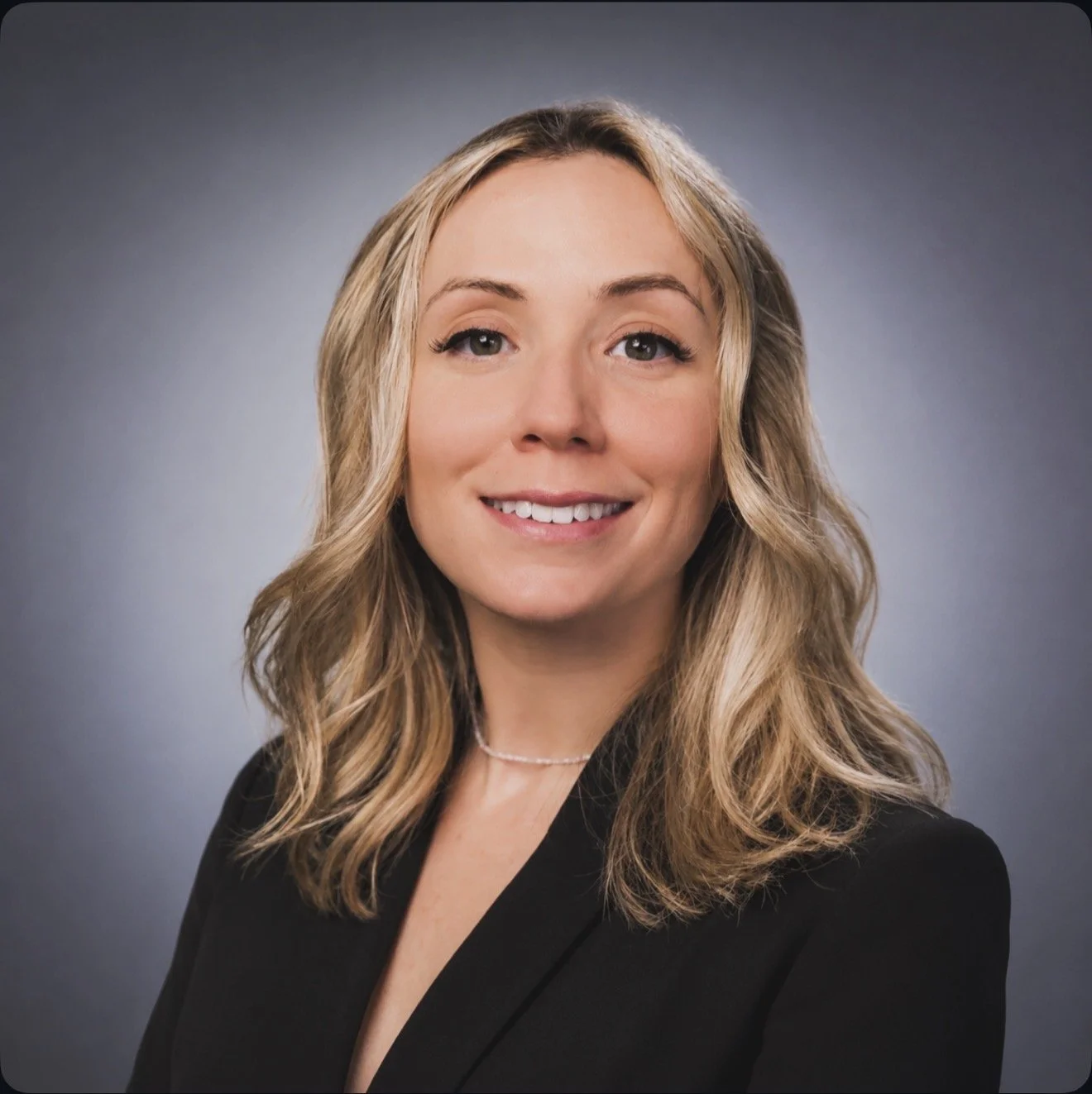 Portrait of a smiling woman with wavy blonde hair, wearing a black blazer and a delicate necklace, against a gray background.