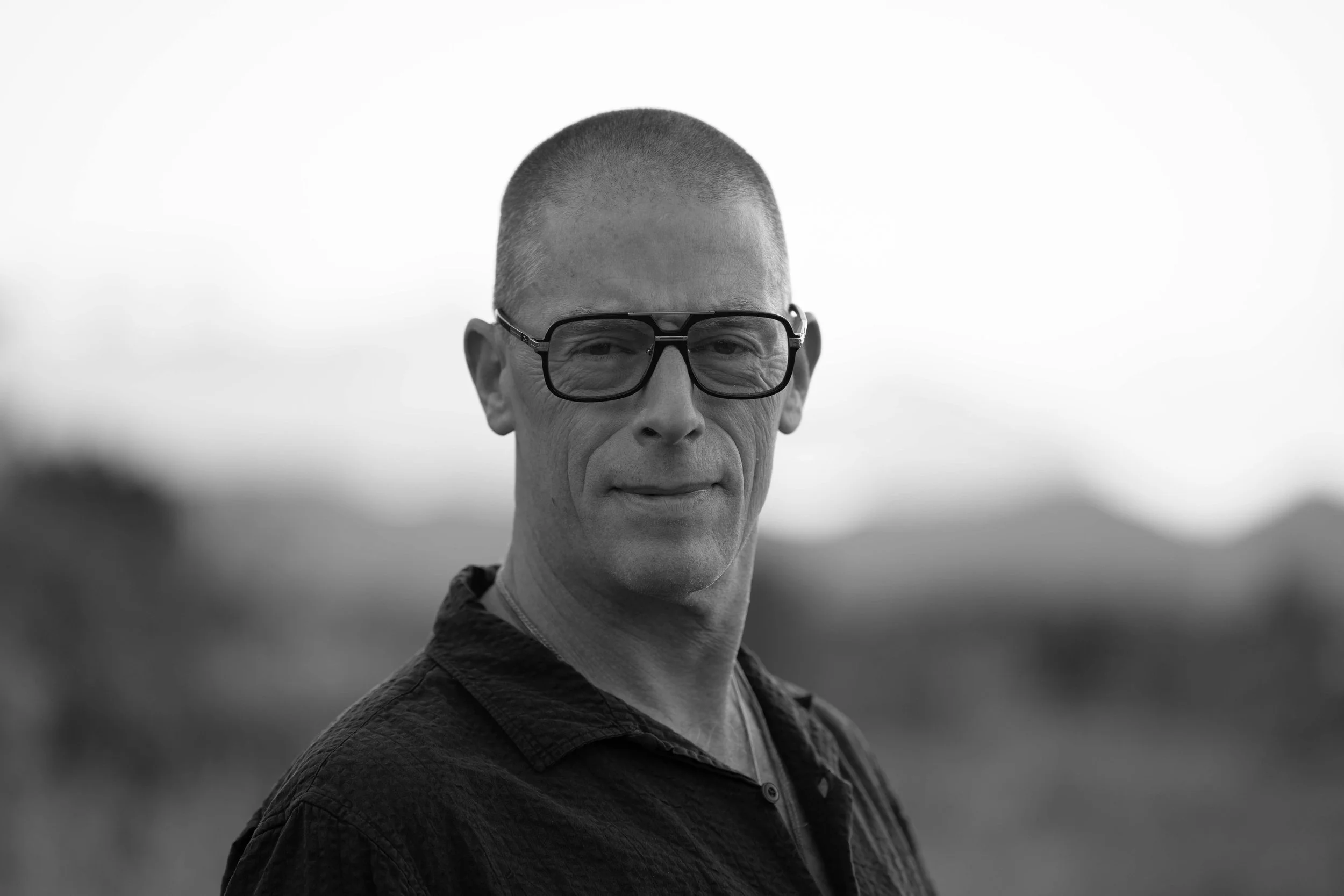 Black and white photo of a middle-aged man with short-cropped hair, wearing glasses and a patterned shirt, standing outdoors with a blurred landscape in the background.
