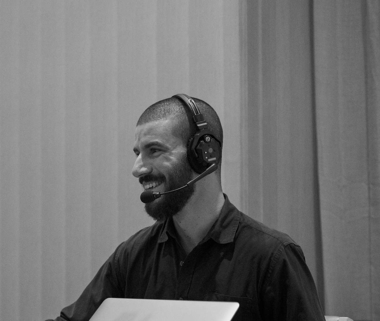 A man with a beard and short hair wearing a headset with a microphone, smiling and sitting in front of a laptop, in a professional setting.