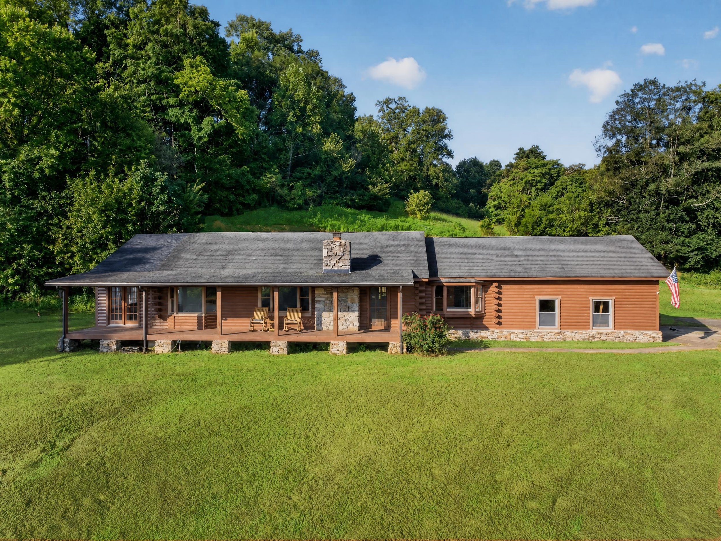 Exterior view of Refuge Ranch with a covered porch, stone chimney, American flag, spacious green lawn, and wooded Tennessee hills in the back ground.