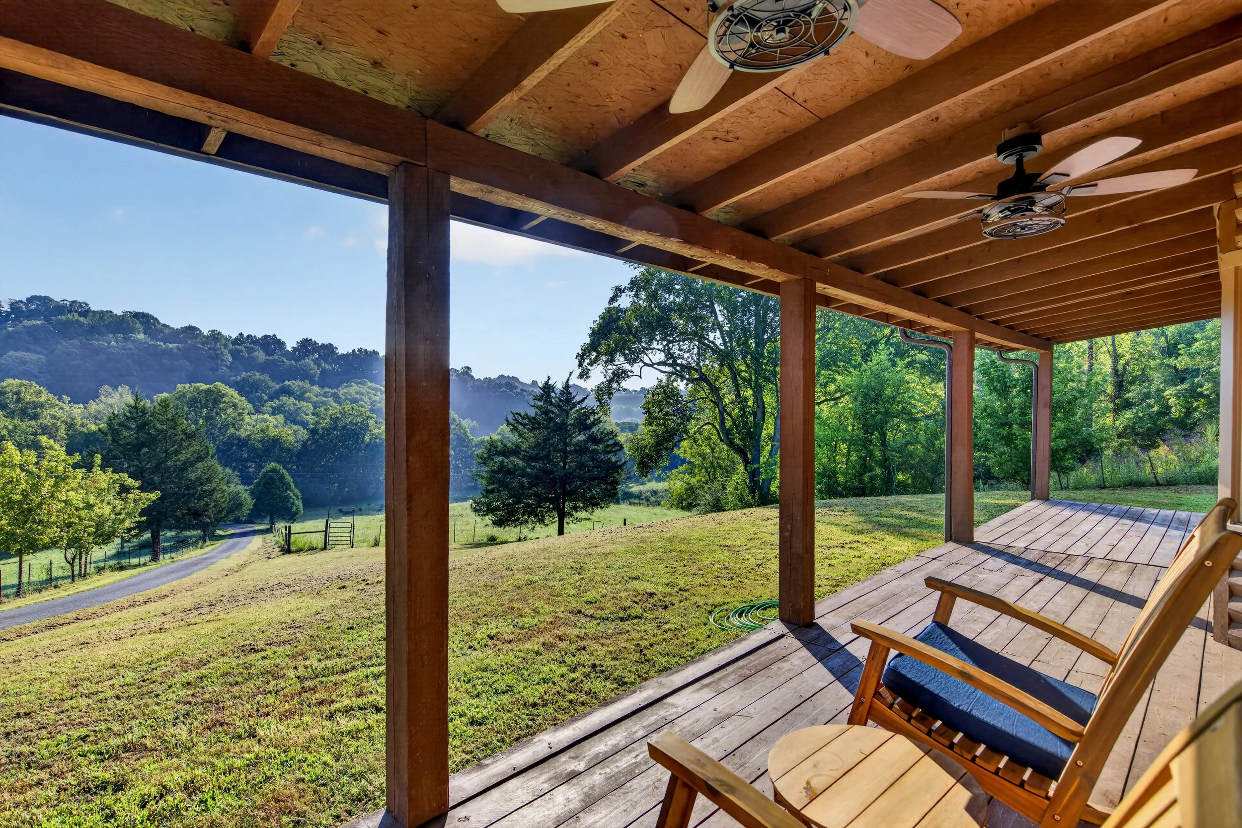 Covered porch at Refuge Ranch with wooden chairs overlooking the green lawn and distant trees, with ceiling fans and outdoor lighting.