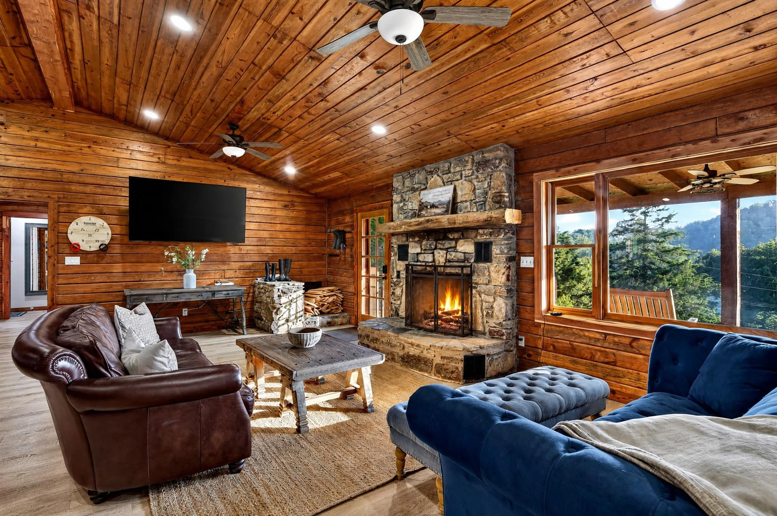 Wood-paneled living room with a stone fireplace, a television, ceiling fans, and large windows overlooking trees.