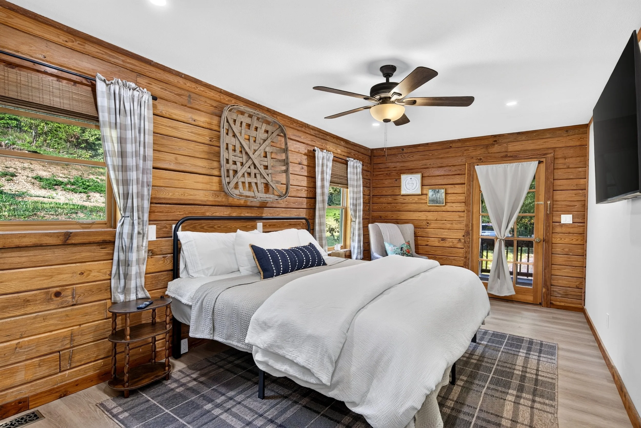 King guest bedroom at Refuge Ranch with white bedding, warm wood-paneled walls, navy accent pillow, wooden nightstand, ceiling fan, and windows with light curtains.
