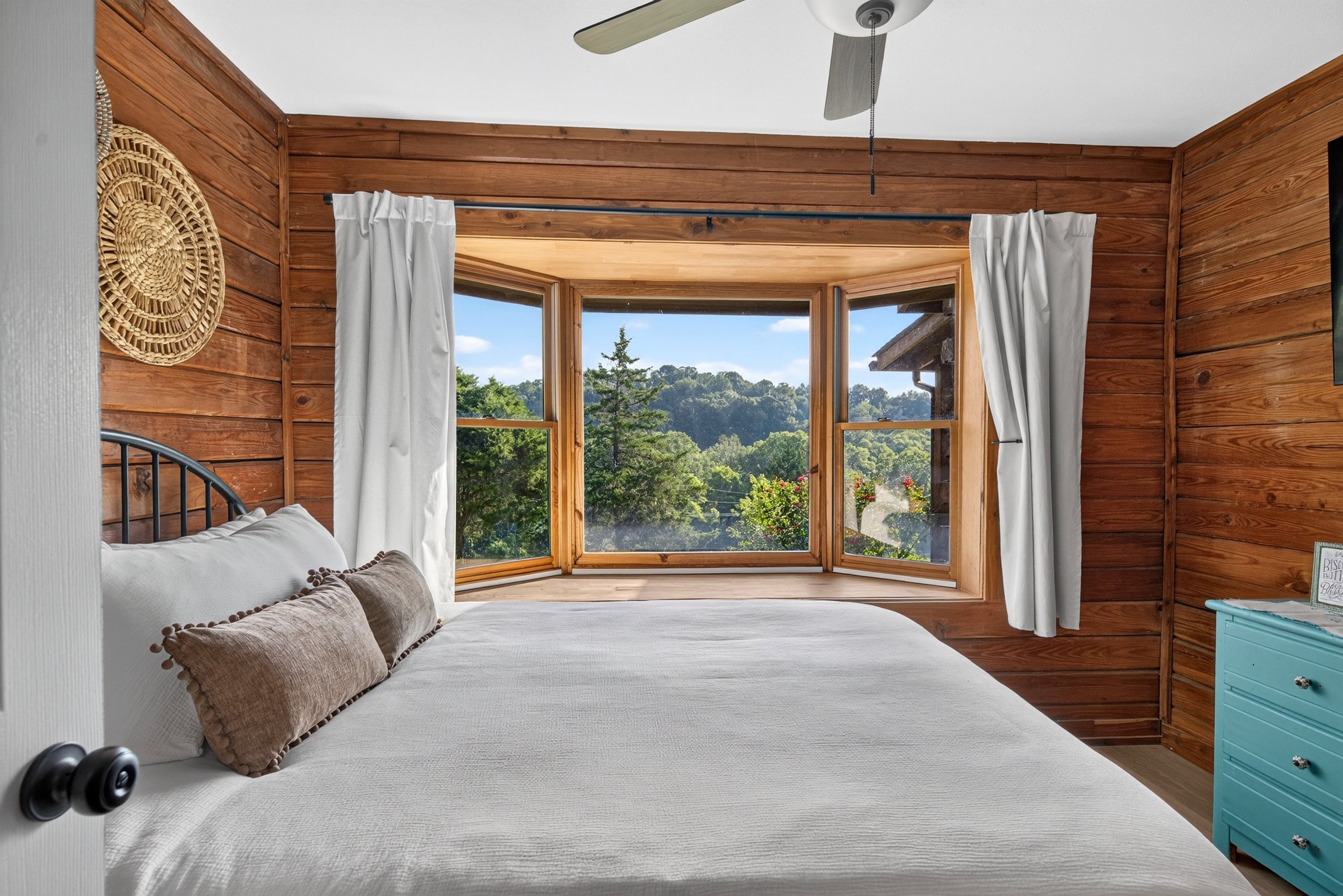 Front guest bedroom at Refuge Ranch with white inviting bedding, warm wood-paneled walls, and a bright window overlooking the trees and sunrise views.