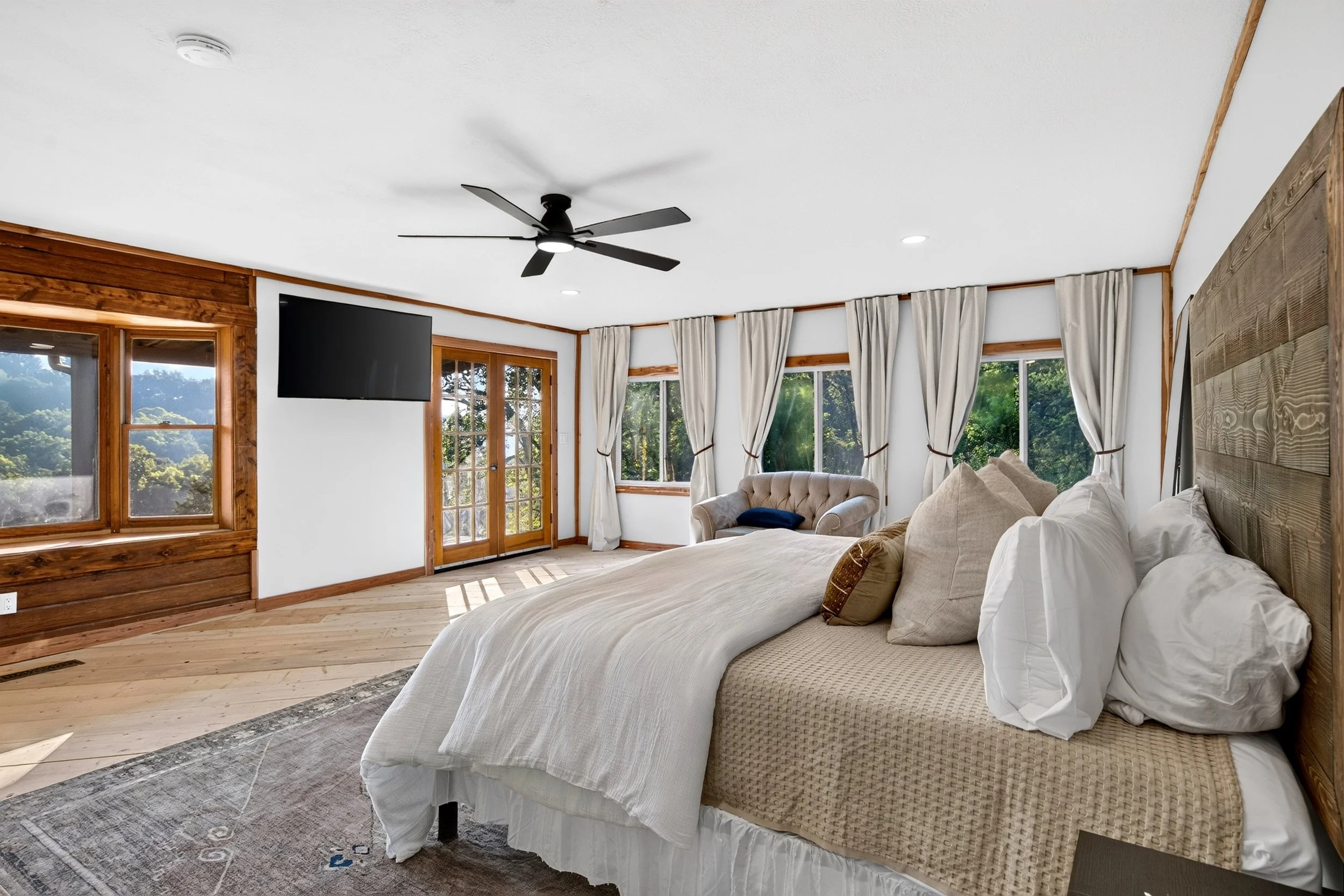 Master suite with large windows, beige curtains, ceiling fan, TV, a bed with beige bedding, and wooden custom headboard with plenty of room.