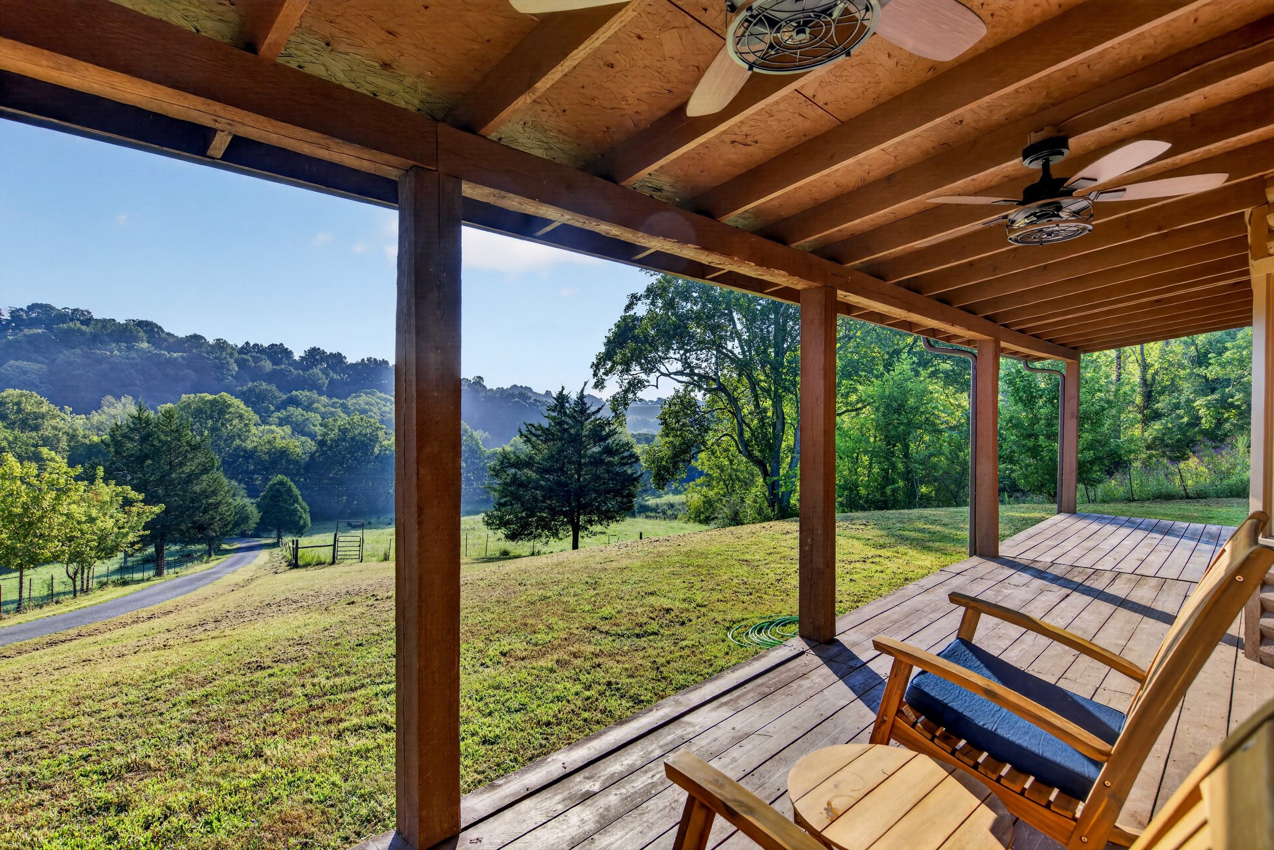 Covered porch at Refuge Ranch overlooking the spacious green lawn, mature trees, and rolling Tennessee hills, with ceiling fans overhead.