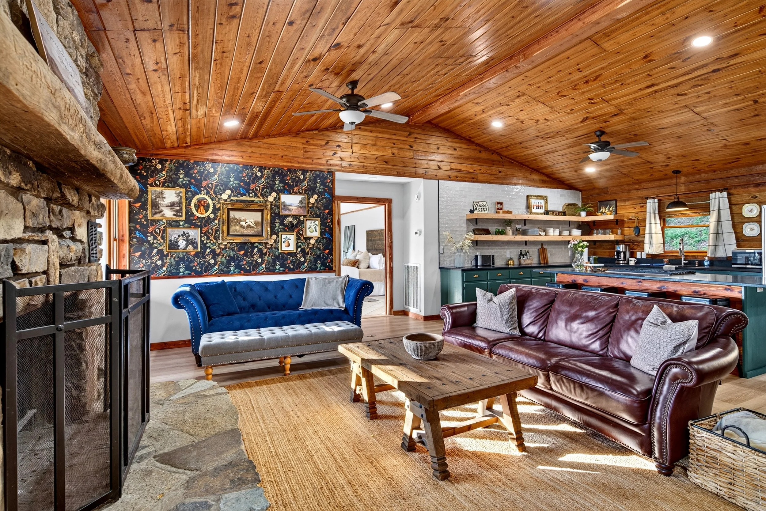 Living room at Refuge Ranch with vaulted wood ceiling, stone fireplace, floral accent wall, comfortable seating and the open-concept kitchen in the background.