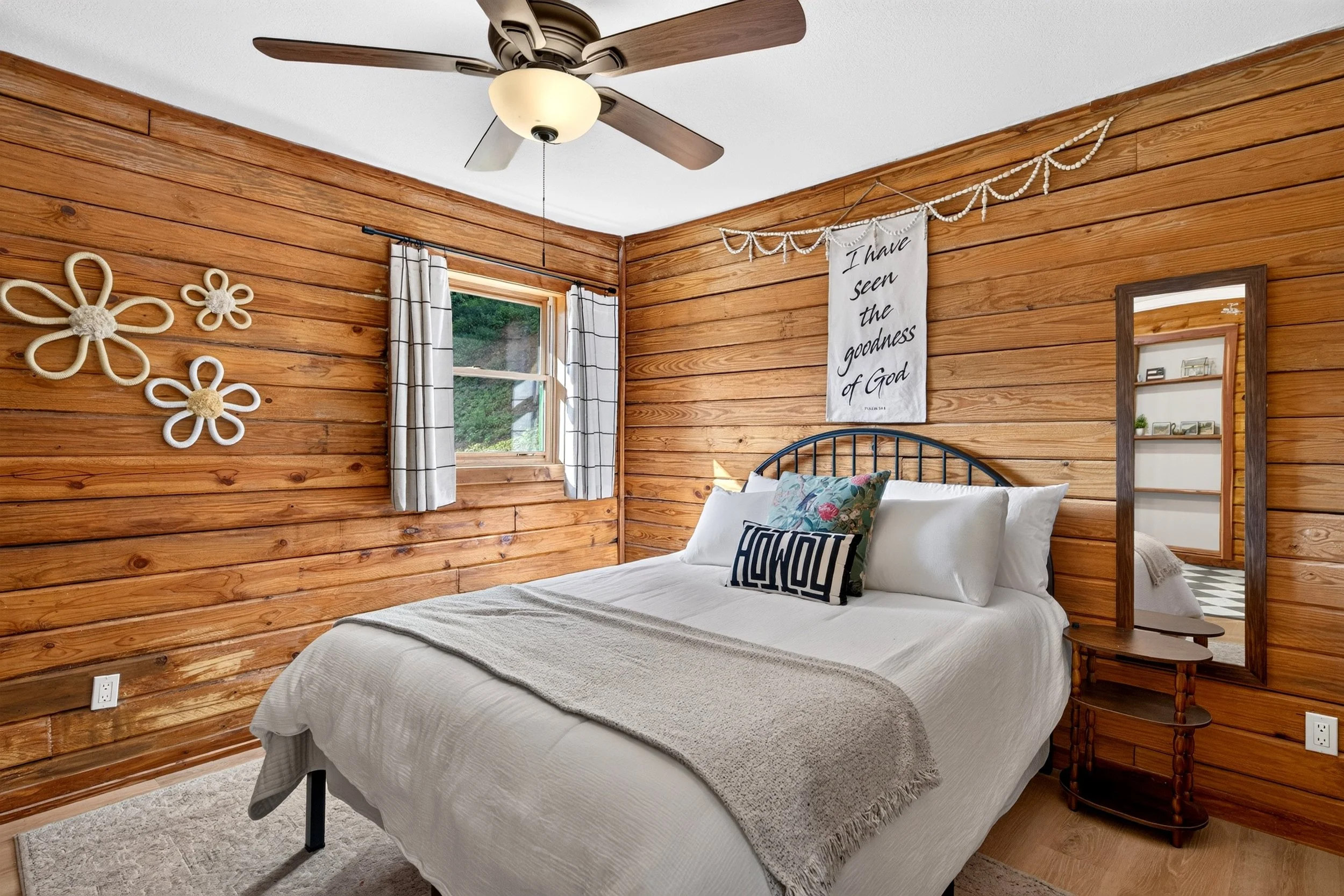 Guest bedroom at Refuge Ranch with white bedding, warm wood-paneled walls, decorative pillows, wall art, ceiling fan, and window with curtains.